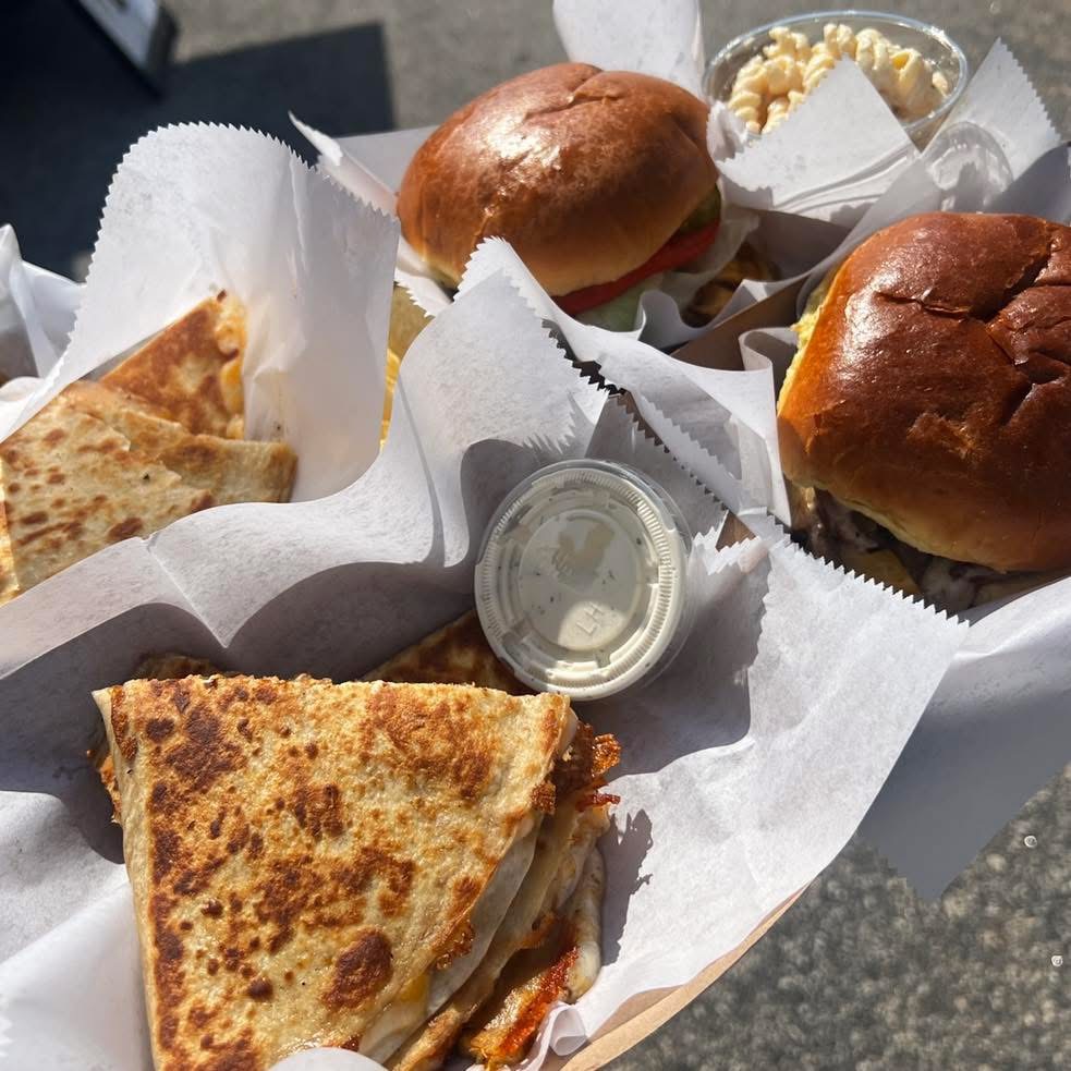 Three sandwiches with bread rolls, a small container of sauce, and a side of potato chips in paper wrappers.