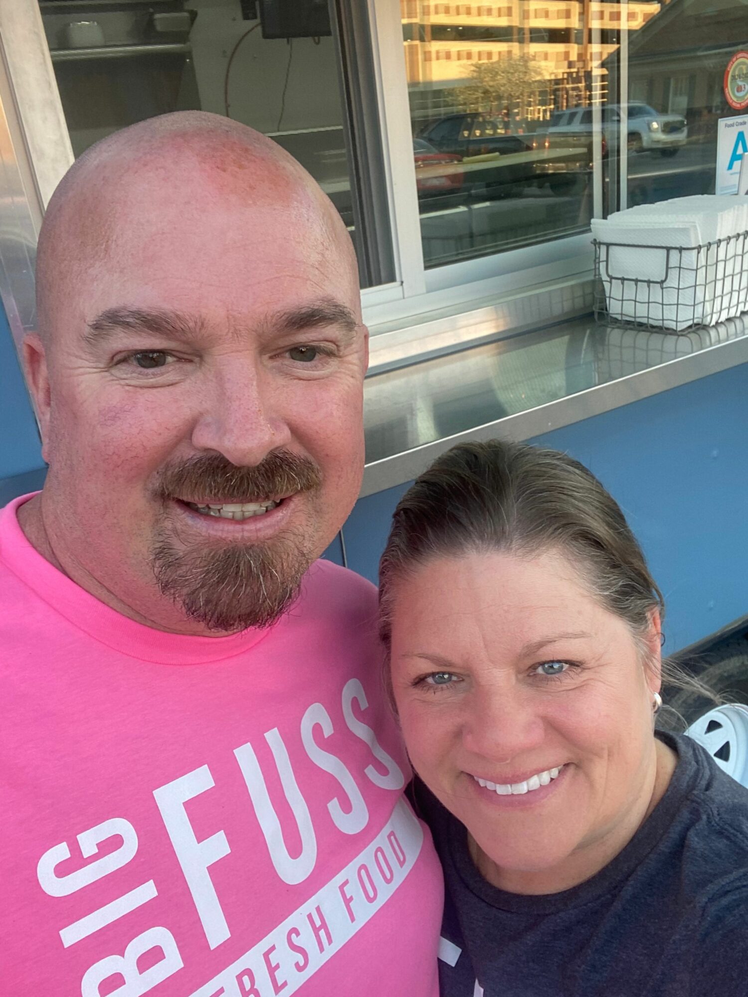 Two smiling people taking a selfie, one with a shaved head and the other with long hair, outside a building.