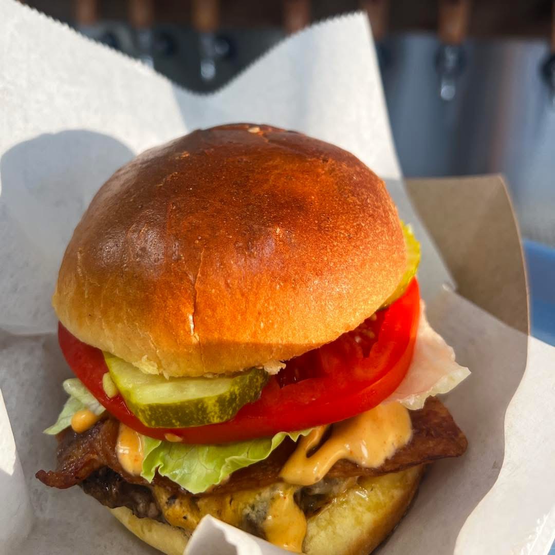 Burger with bun, lettuce, tomato, pickles, cheese, and meat patty, served in a paper wrapper.