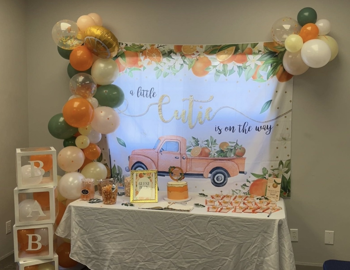 Decorative table with balloons, a pink truck, and a backdrop with floral design and text for a baby shower.