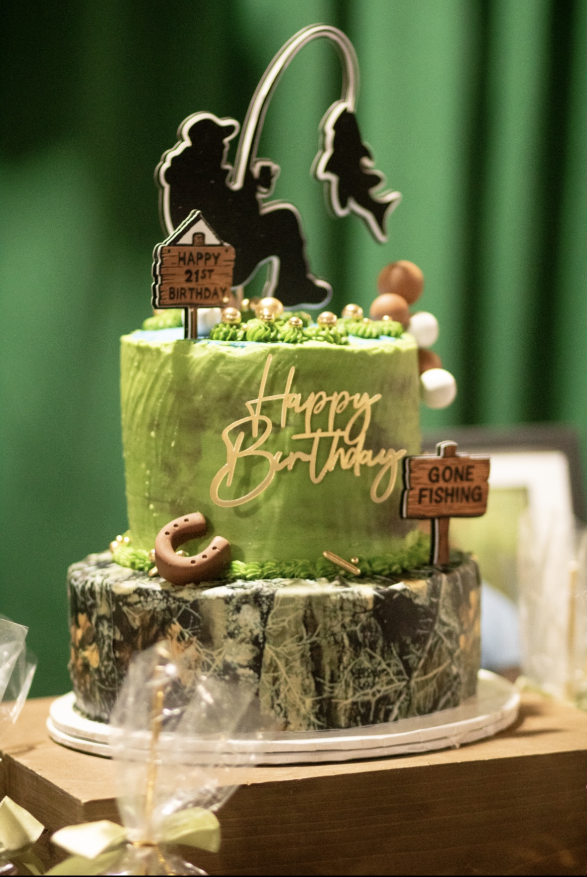 Two-tiered birthday cake with fishing-themed decorations, green and marbled bases, and a fishing rod topper, on a wooden table.