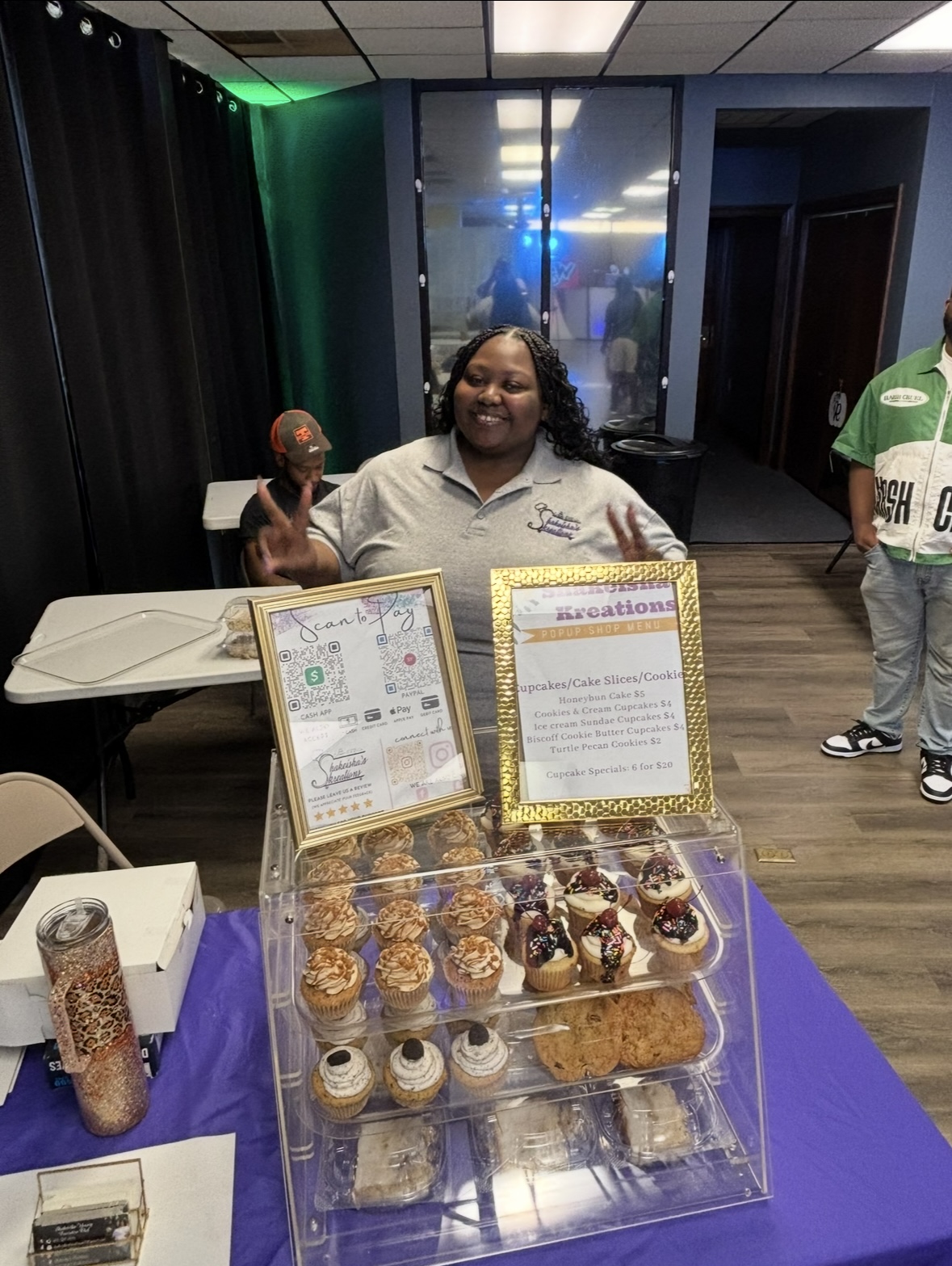 Smiling woman behind a table with cupcakes and desserts, holding a cupcake, in an indoor event space.
