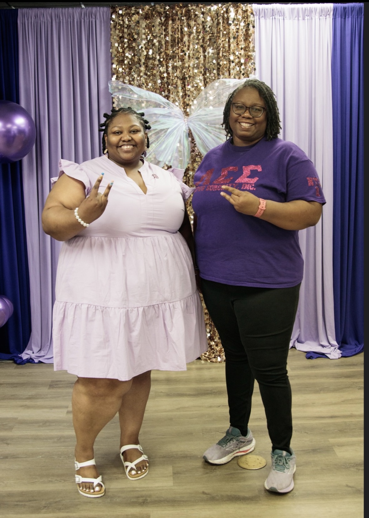 Two women smiling and posing in front of a decorated backdrop with purple and gold curtains, one making a peace sign.