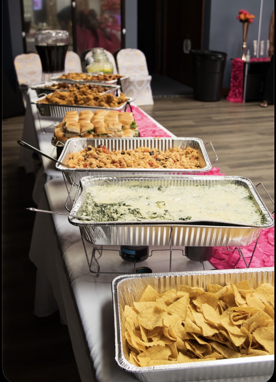 Buffet table with various dishes in aluminum trays, including chips, pasta, and baked items, in a decorated room.