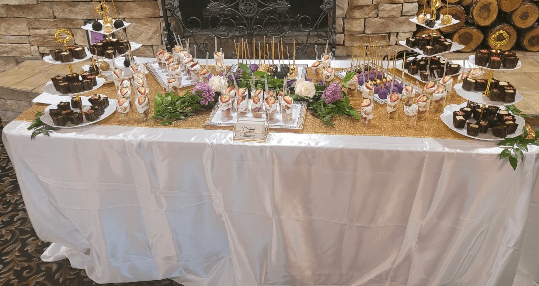 Decorated table with desserts, treats, and floral arrangements, covered with a white tablecloth, in front of a brick wall.