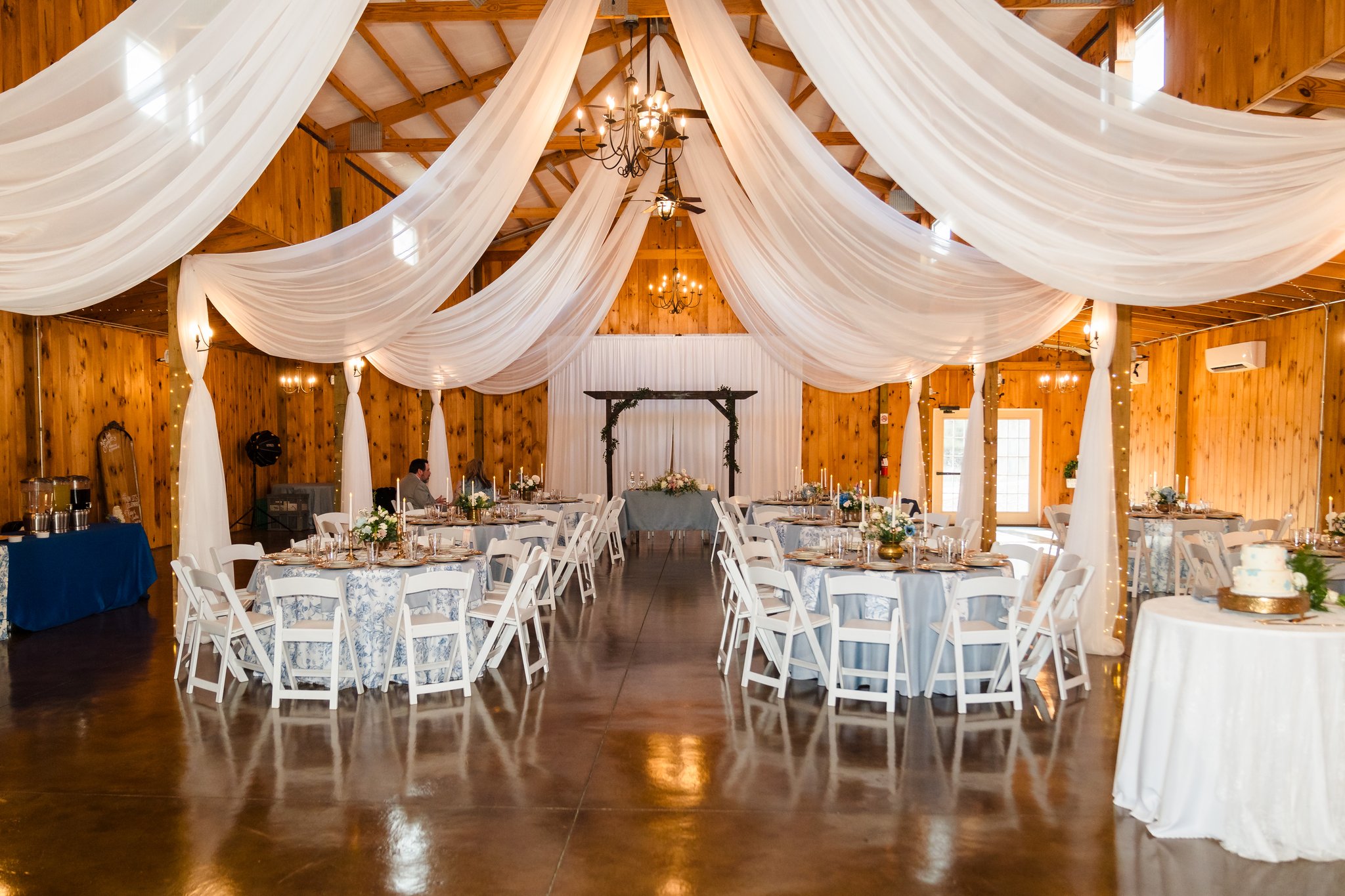 Elegant event space with draped white fabric ceiling, wooden walls, round tables with white chairs, and a chandelier, set for a celebration.