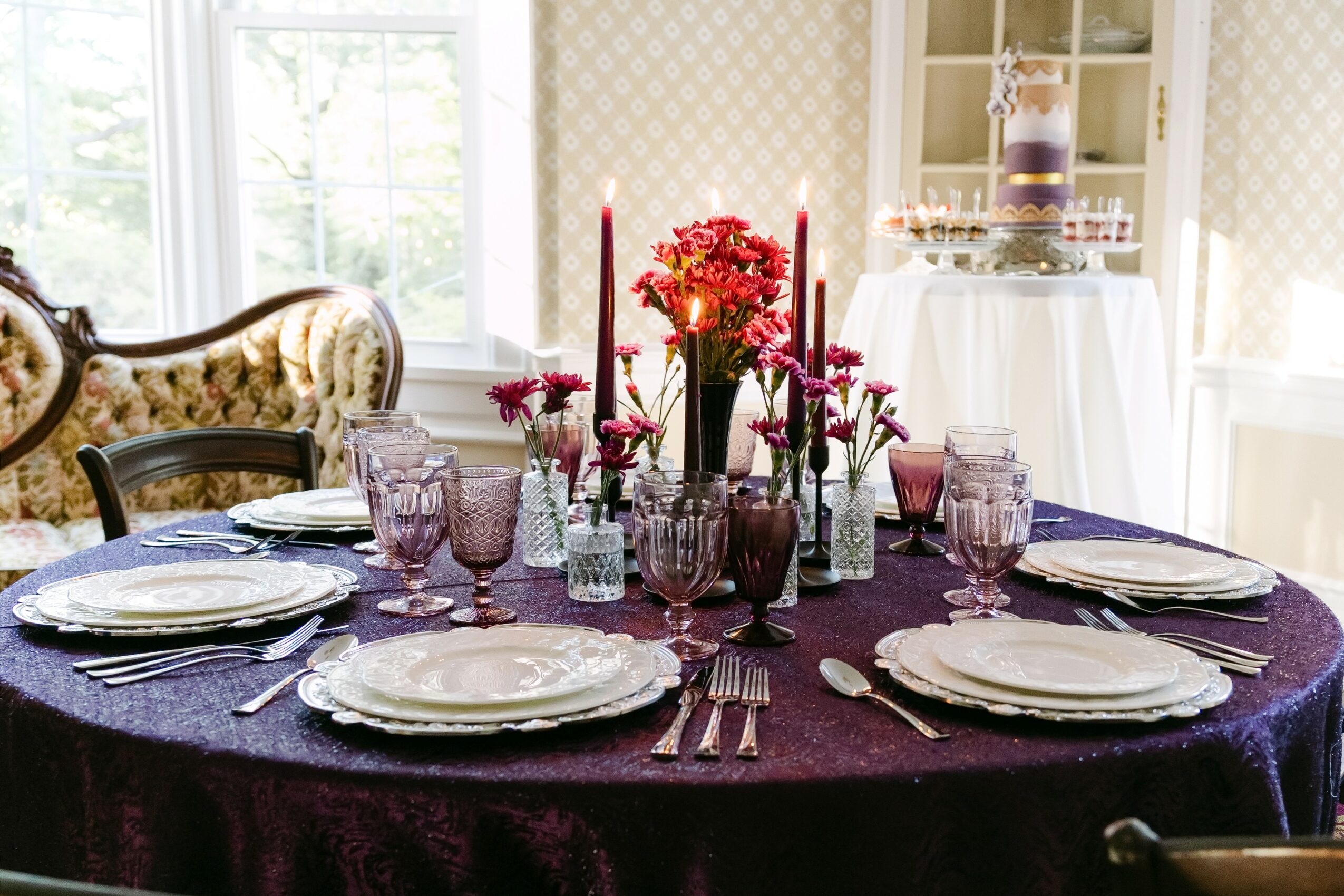 Round table with a dark purple tablecloth, set with plates, glasses, and silverware, decorated with pink and red flowers and tall candles.