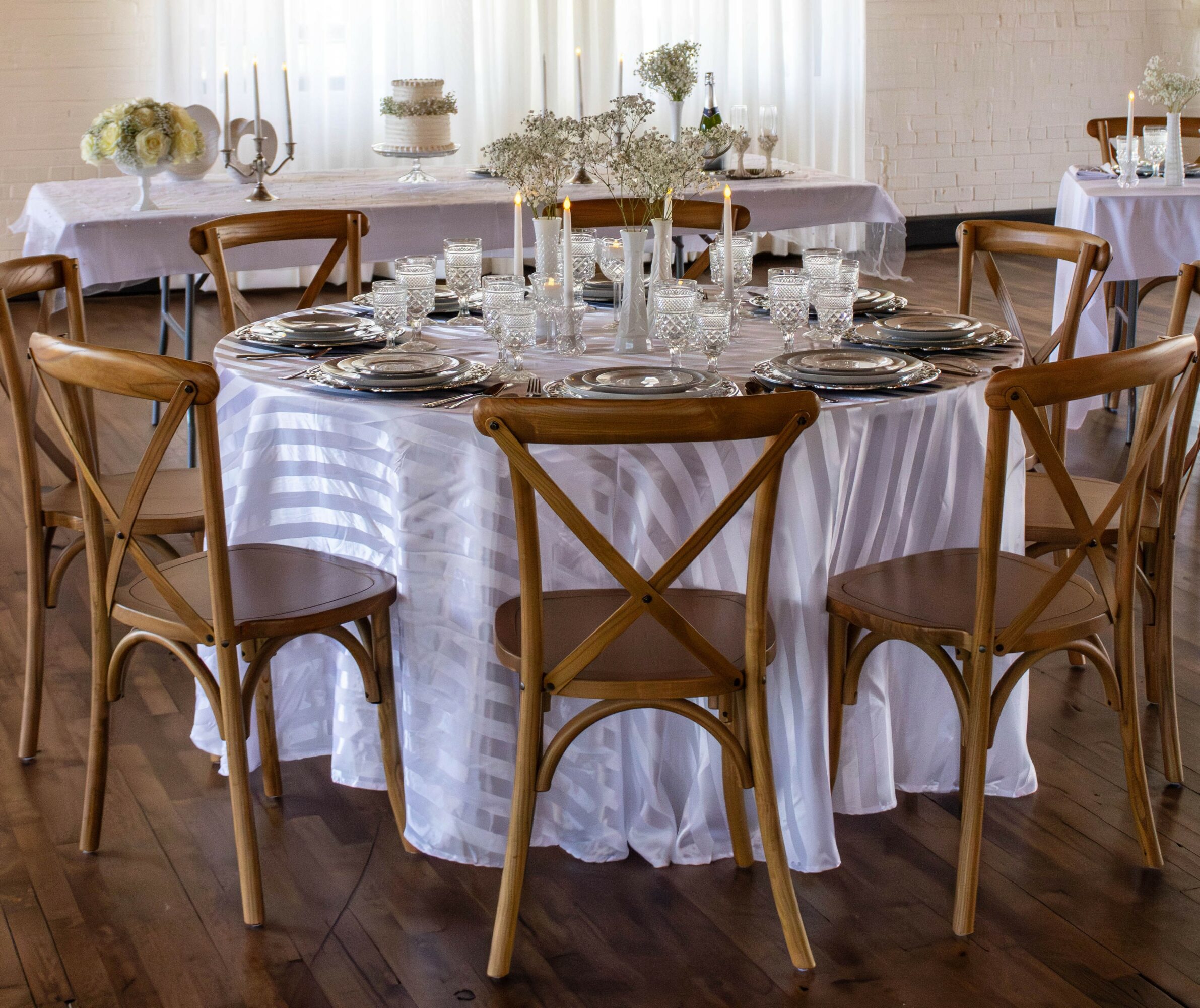 Round table with white tablecloth, surrounded by wooden chairs, set with glassware and plates, in a bright room.