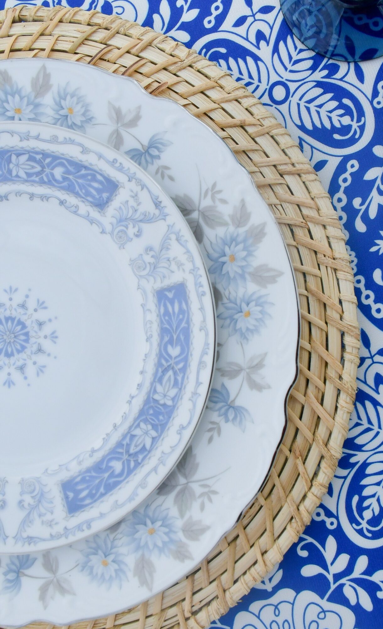Stacked plates with blue and white patterns on a woven placemat, on a blue patterned tablecloth.