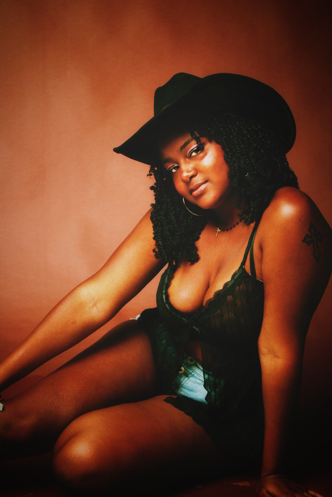 Woman wearing a black hat and lace top, sitting with one arm extended, against a warm brown background.