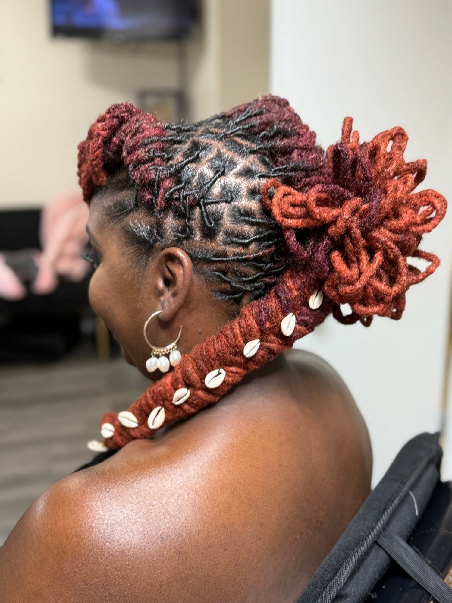 Side view of a woman with styled dreadlocks in red and black, decorated with white shells, in a salon setting.