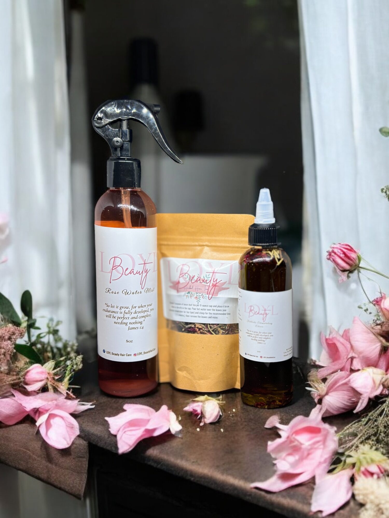 Three brown bottles with labels, surrounded by pink flowers on a dark surface, with a window and curtain in the background.