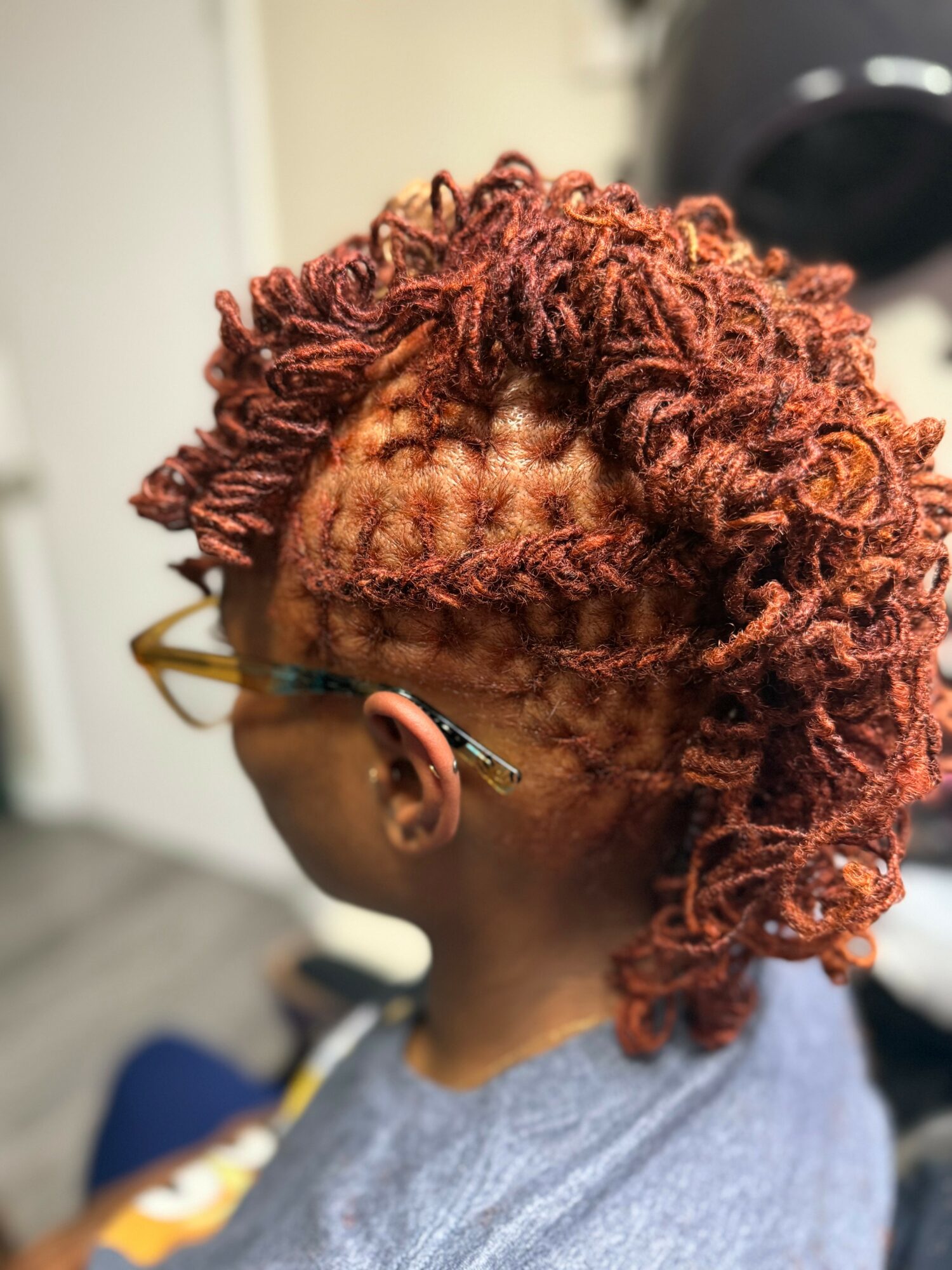 Side view of a person with curly, reddish-brown hair styled in a mohawk, wearing glasses and a gray shirt.