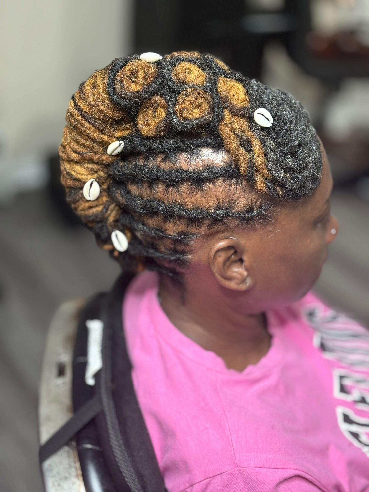 Side view of a young girl with styled, decorated dreadlocks adorned with white beads, wearing a pink shirt.