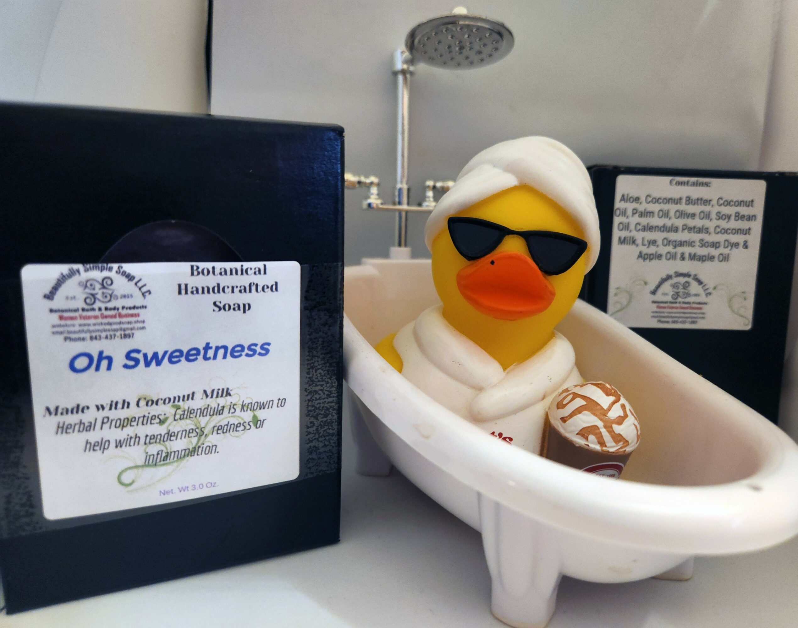 Rubber duck wearing sunglasses in a bathtub with soap, next to a soap box labeled 'Oh Sweetness' and other soap boxes.