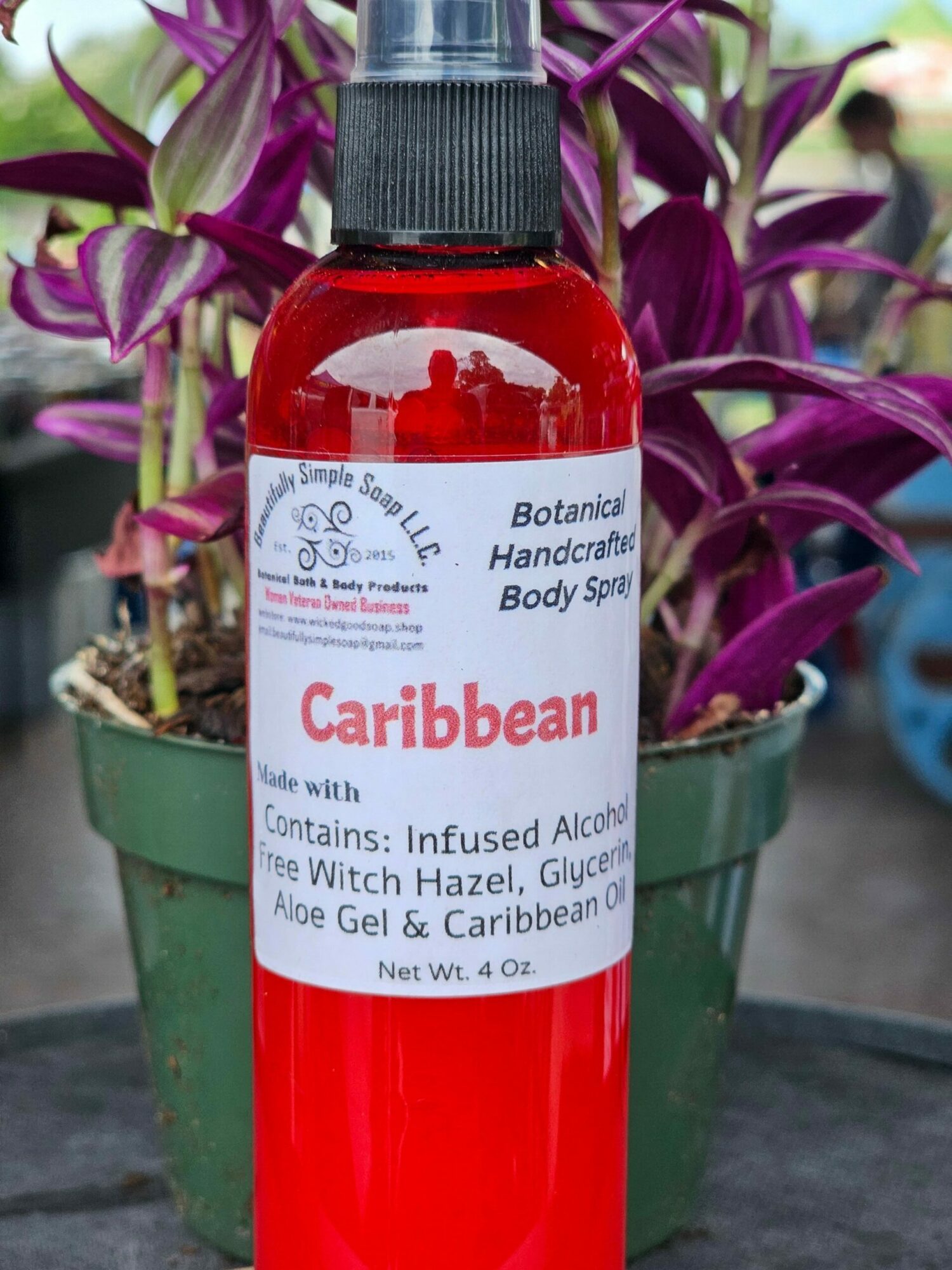 Red bottle of Caribbean body oil with a white label, placed in a green pot with purple and white flowers.