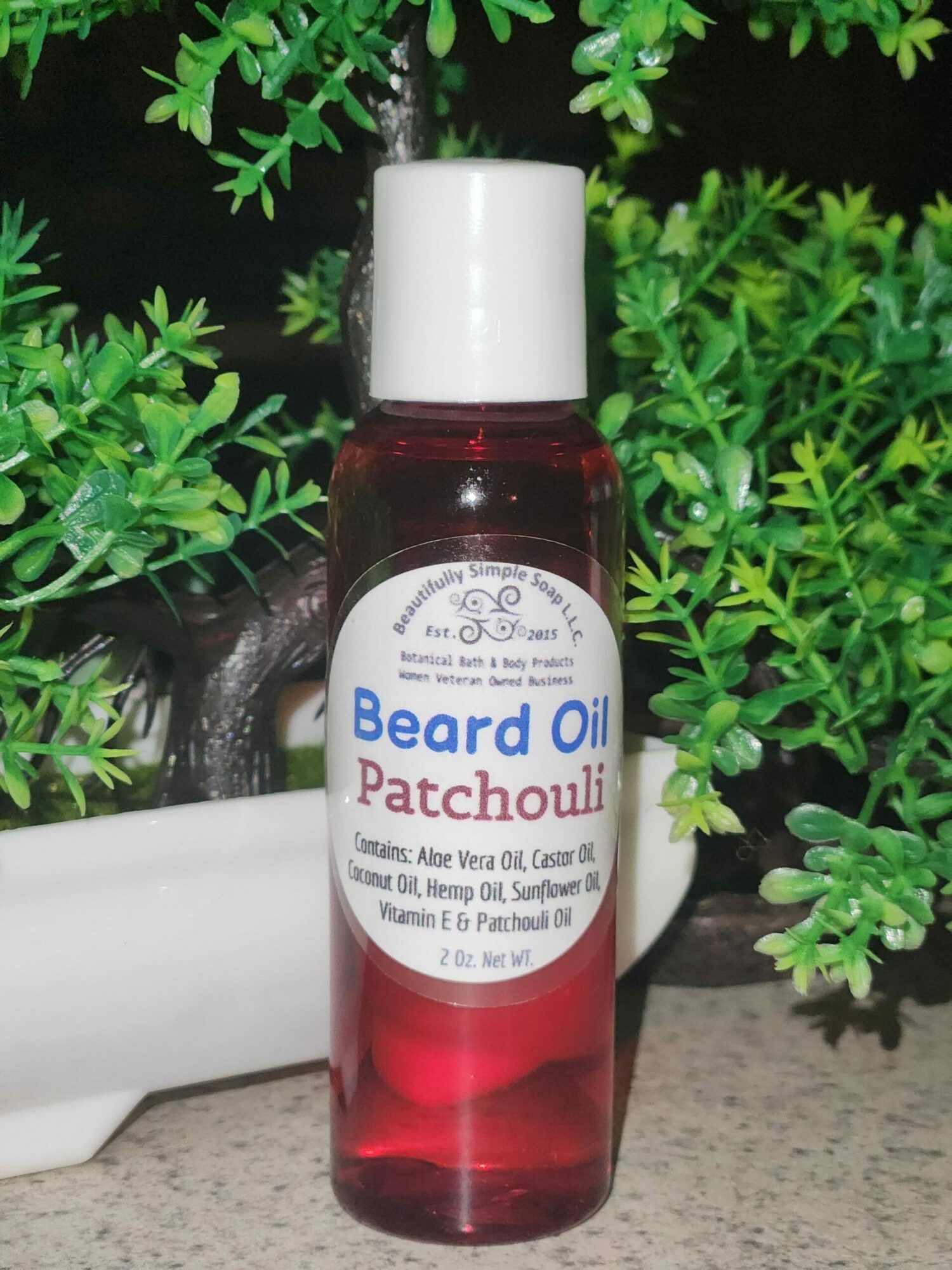 Red bottle of beard oil with white cap, surrounded by green plants, on a gray surface.