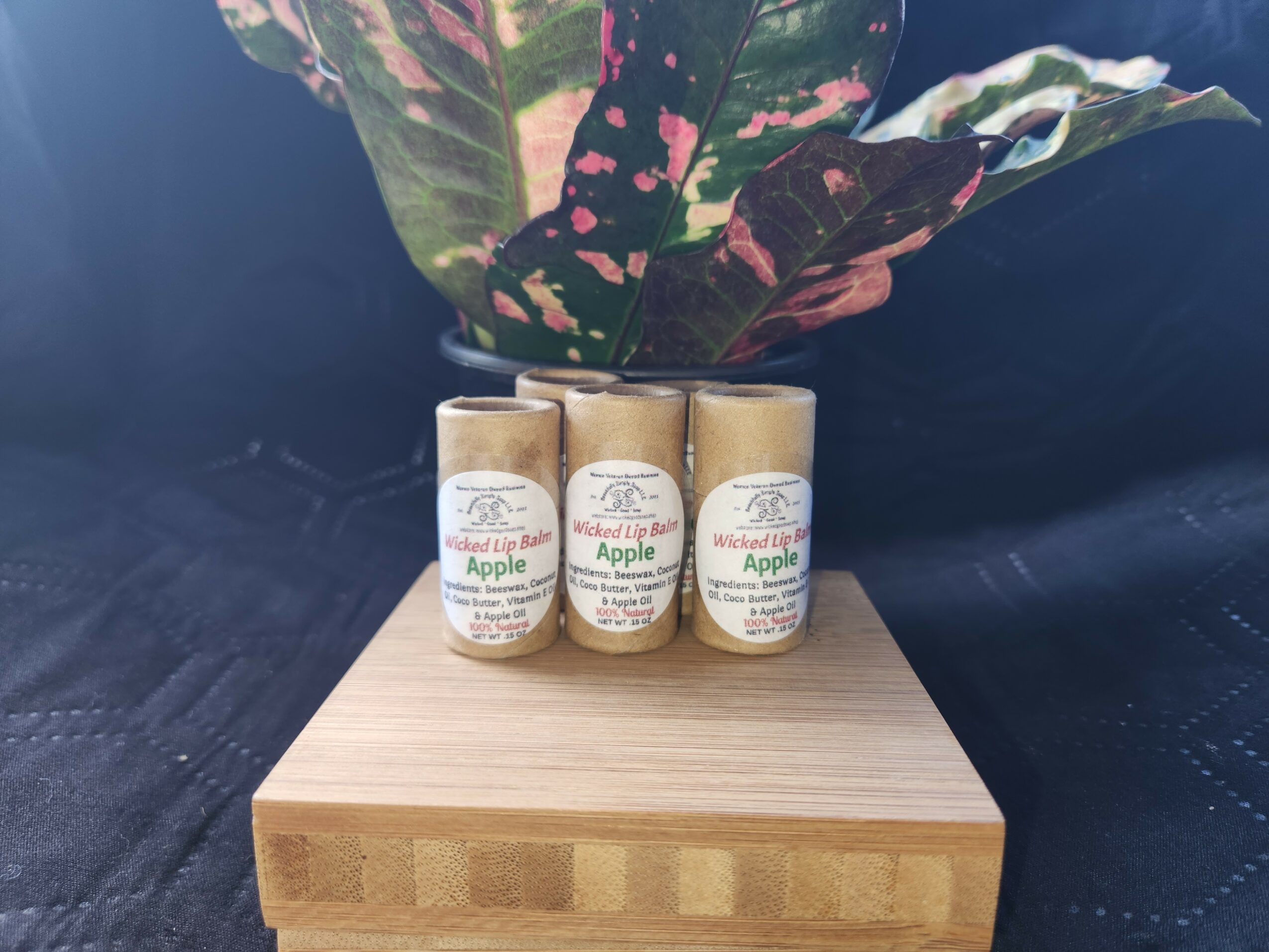 Three small bottles of apple-flavored product in front of a potted plant with variegated leaves on a wooden surface.