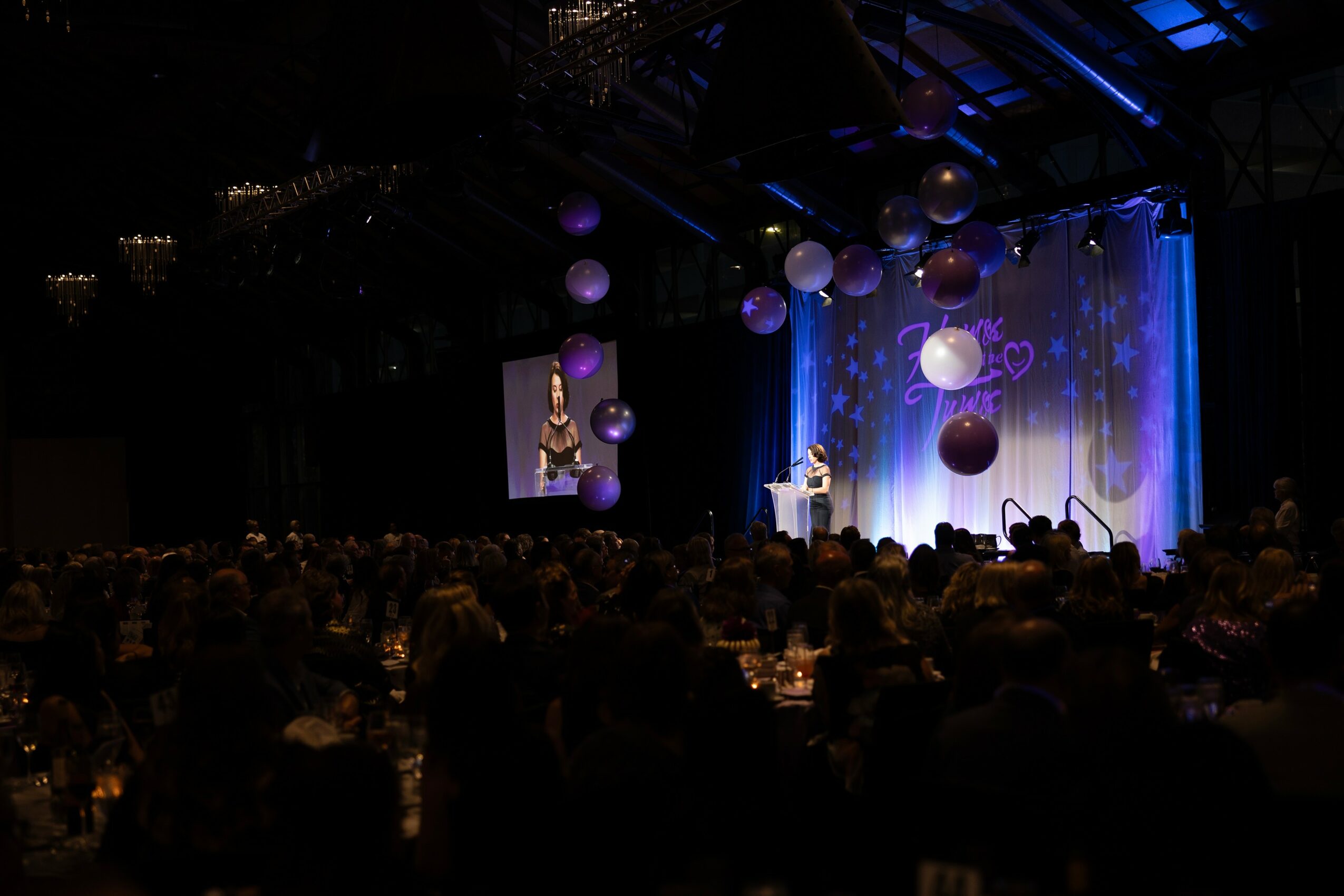 Stage with a speaker, decorated with hanging balloons and blue lighting, audience seated in a darkened room.