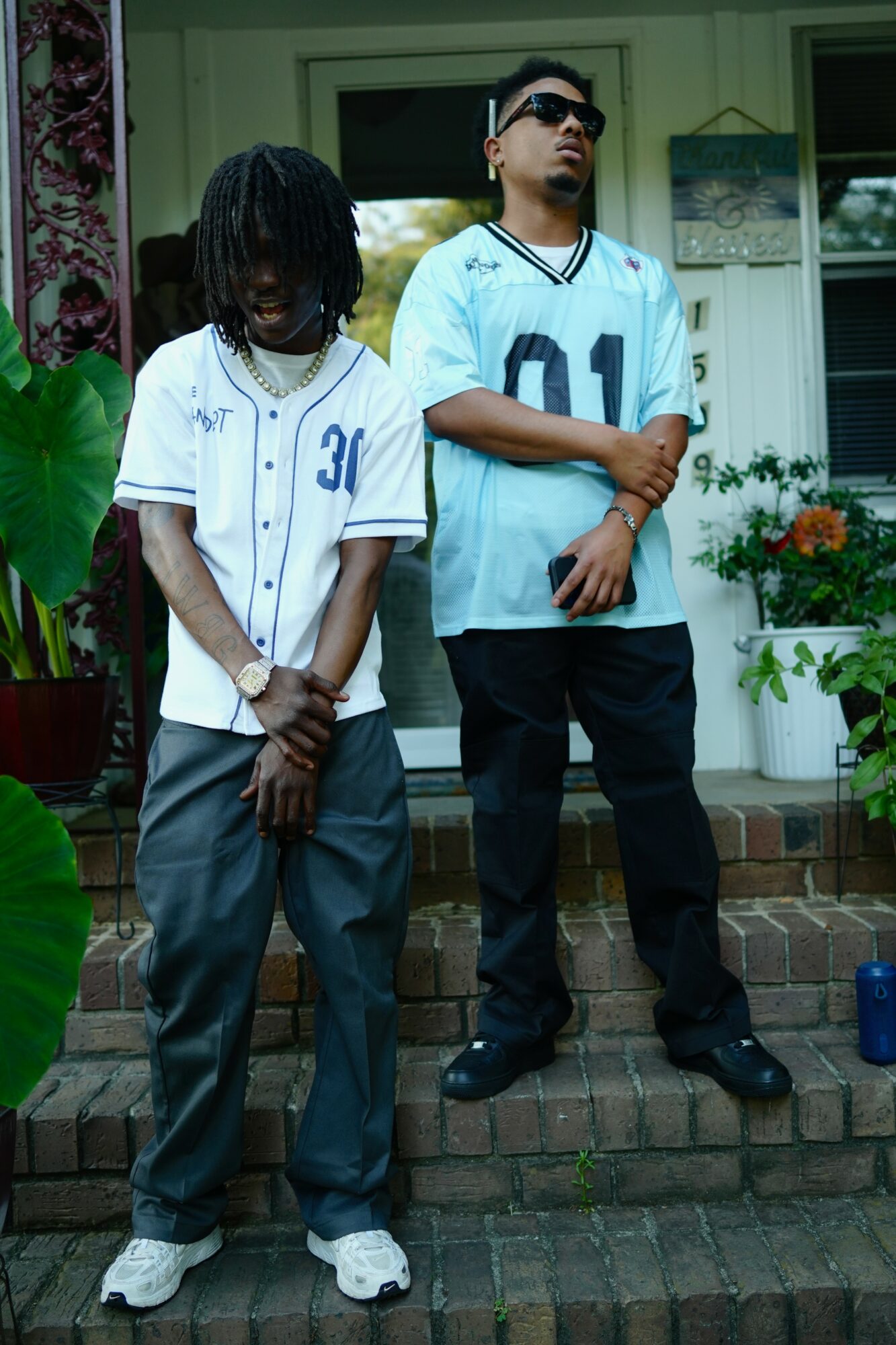 Two young men stand on a brick porch in front of a house, wearing casual clothing and sunglasses.