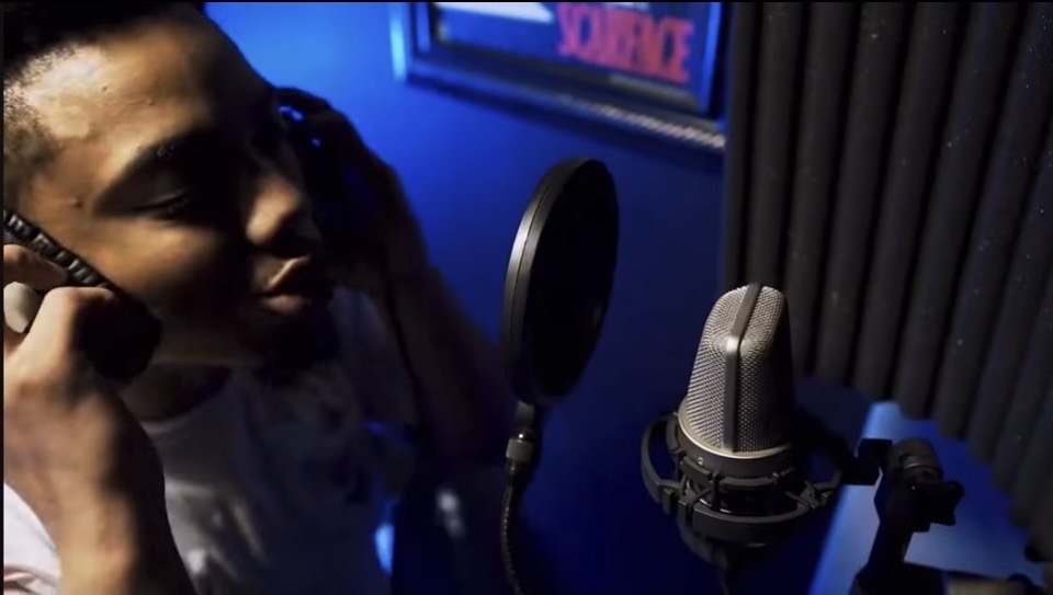 Person recording vocals in a studio with a microphone and pop filter, blue lighting, and soundproofing panels.