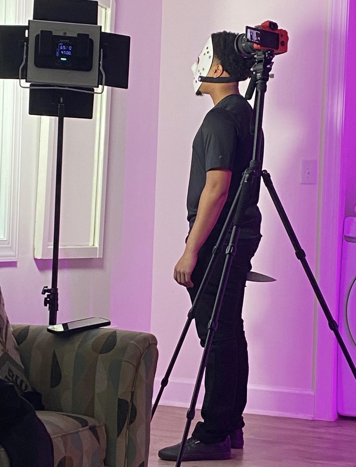 Person wearing a virtual reality headset and standing next to camera equipment in a room with purple lighting.