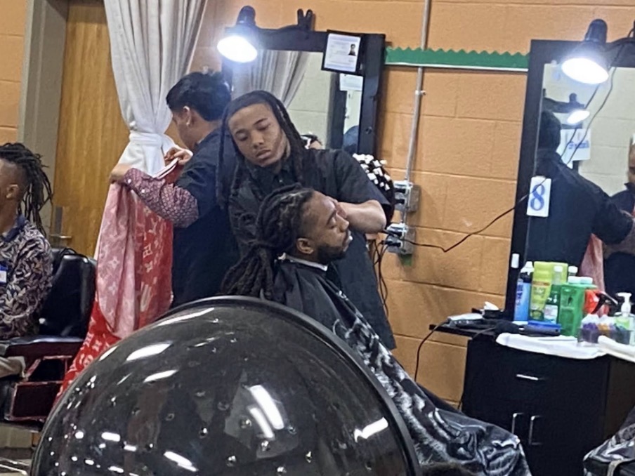 People in a barbershop, one person getting a haircut, mirrors, and hair products visible.