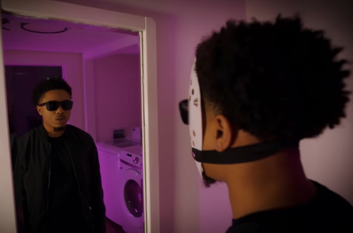 Person wearing a mask facing another person in a room with pink lighting, wearing sunglasses and dark clothing.