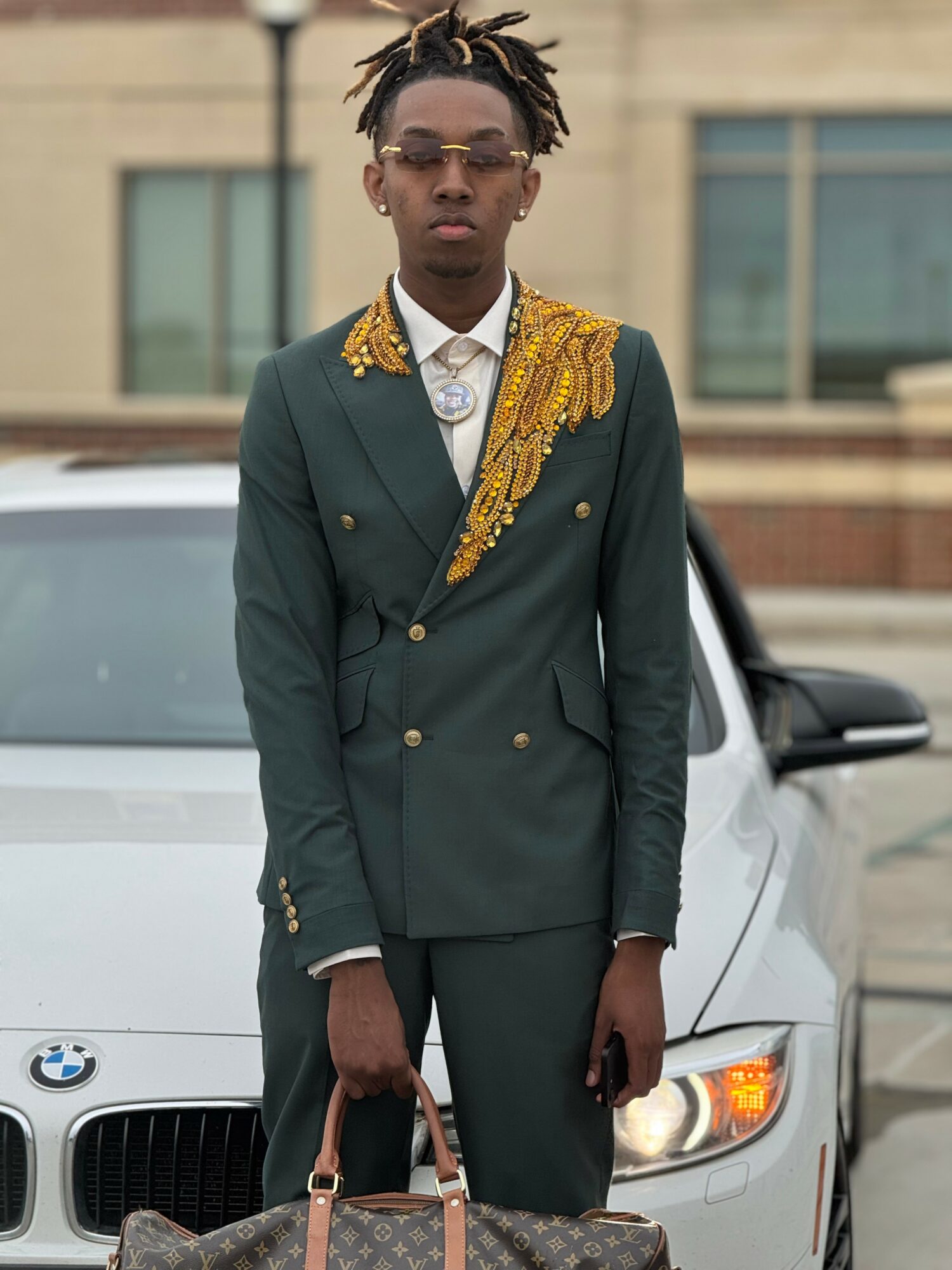 Person in a dark green suit with gold embellishments, standing in front of a white car, holding a handbag.