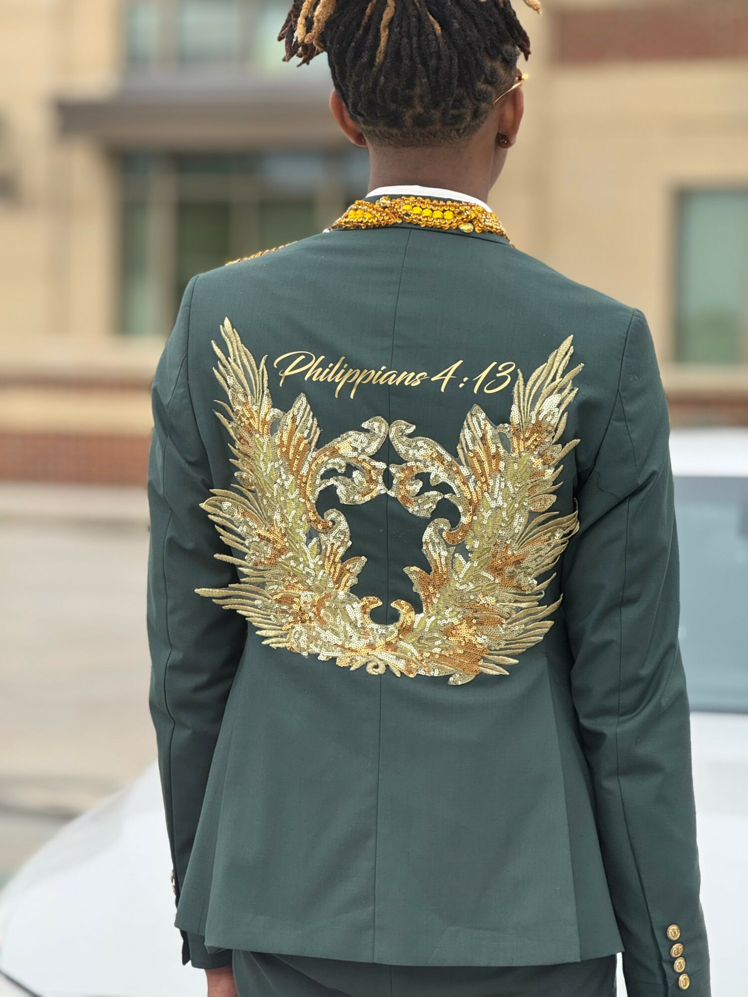 Person with short hair wearing a dark green jacket with gold embroidery and text on the back, outdoors.