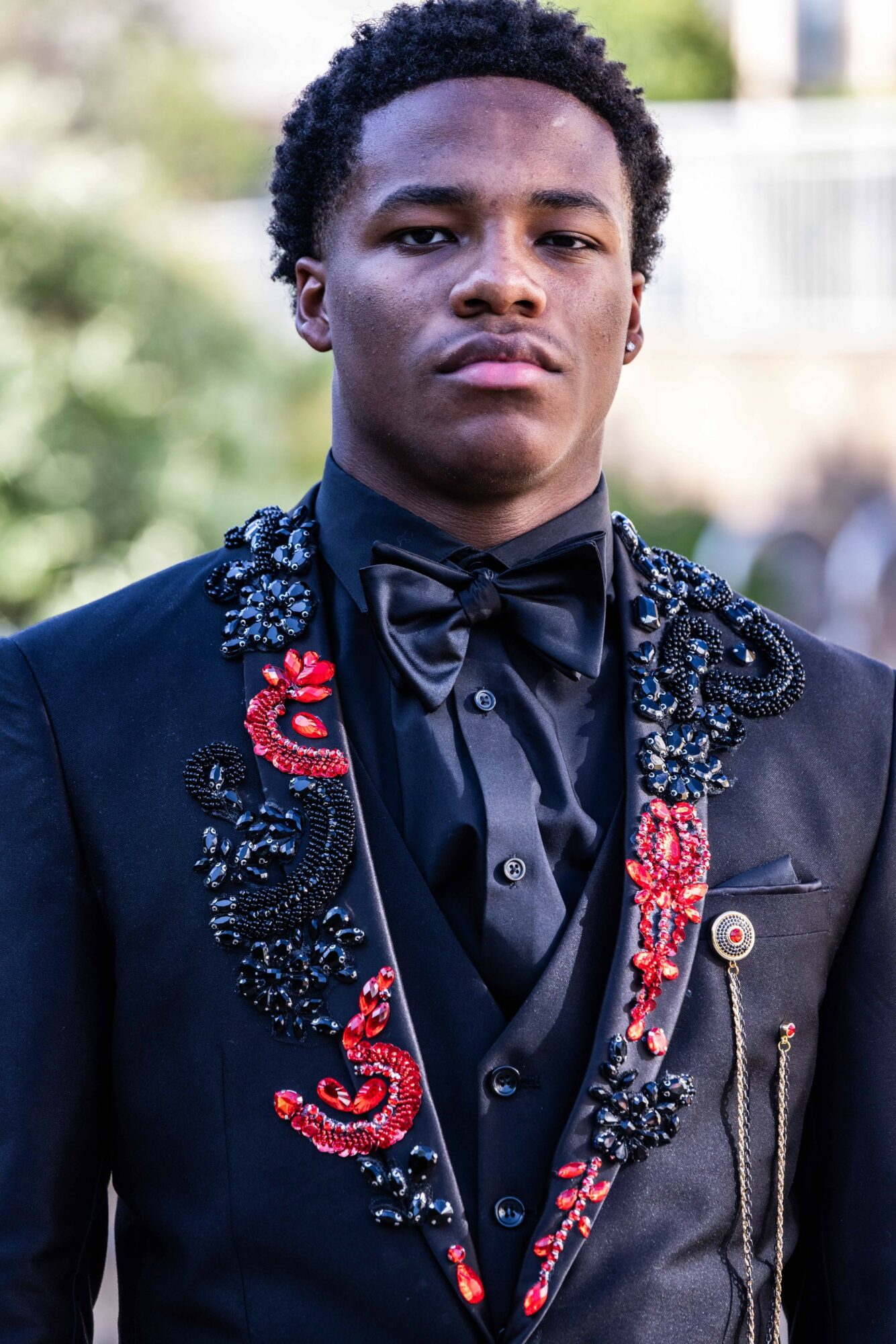 Young man in formal black suit with embroidered details, wearing a bow tie, outdoors with blurred background
