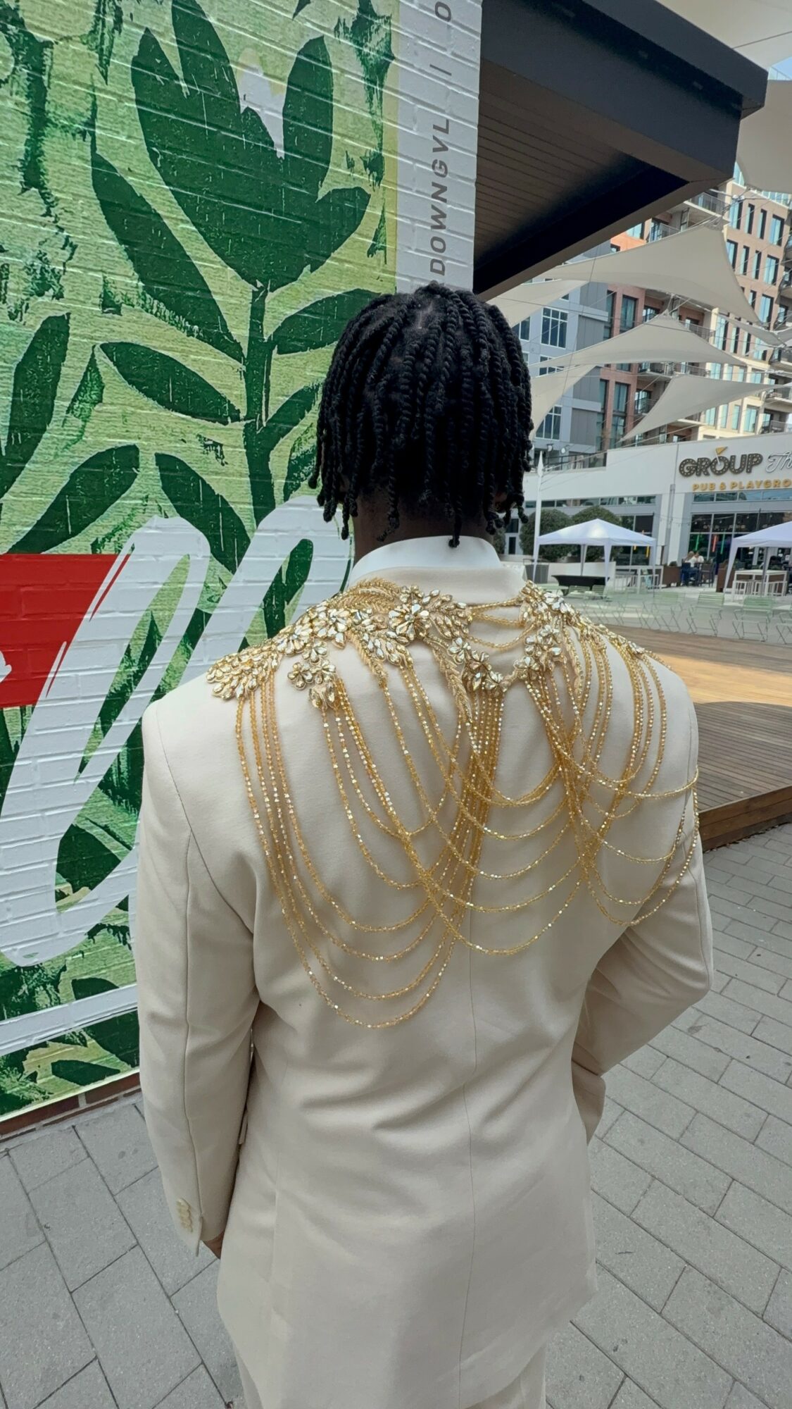 Person with short dreadlocks wearing a white jacket with gold chains on the back, standing outdoors near a green mural.