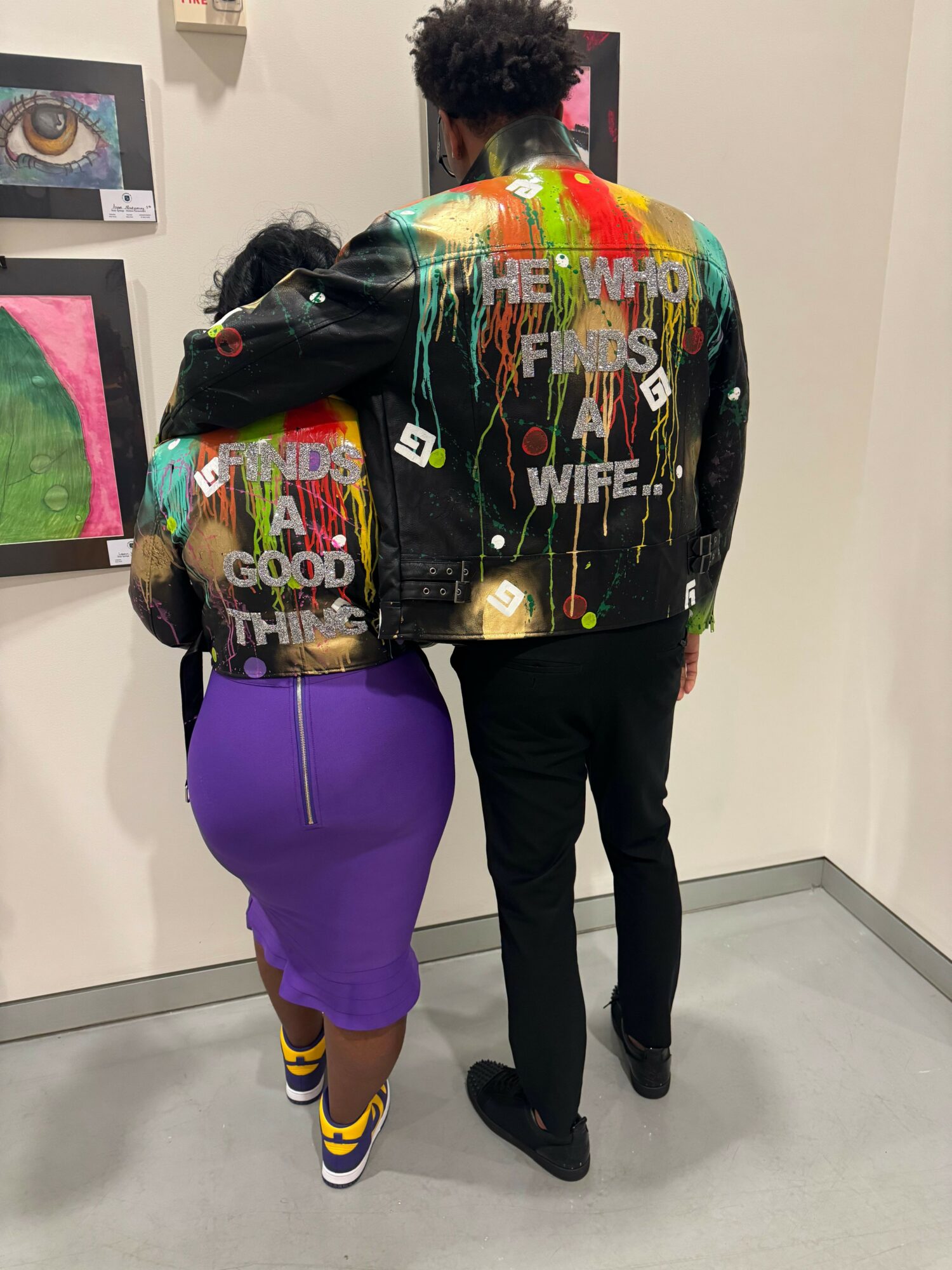 Two people standing in a room, one wearing a colorful jacket and the other in a purple dress and yellow shoes.