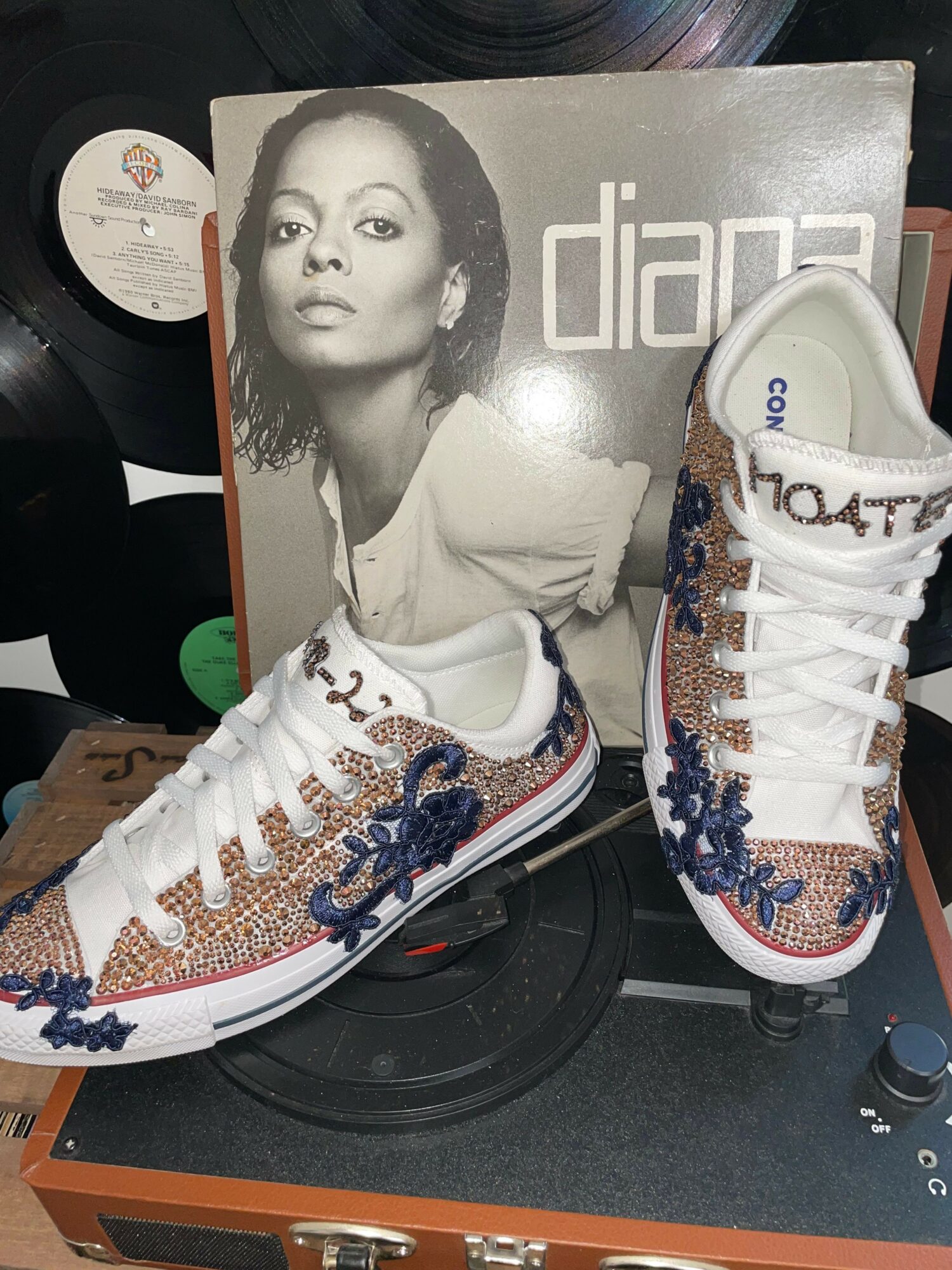 Sneaker with colorful embellishments resting on a vinyl record player, with a black-and-white magazine cover featuring a woman in the background.