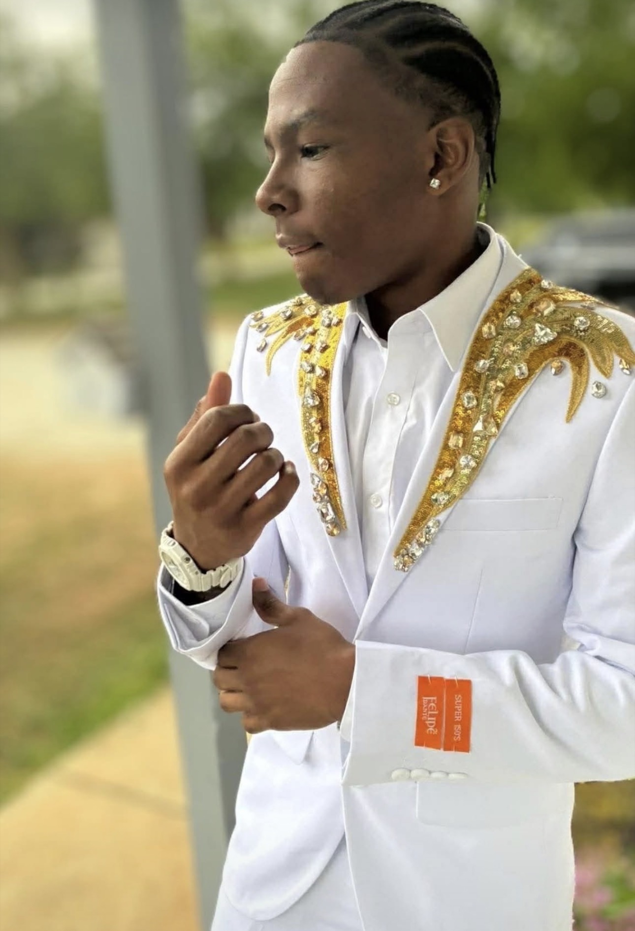 Young person in white jacket with gold embellishments, looking down thoughtfully outdoors, with blurred background of trees and grass.