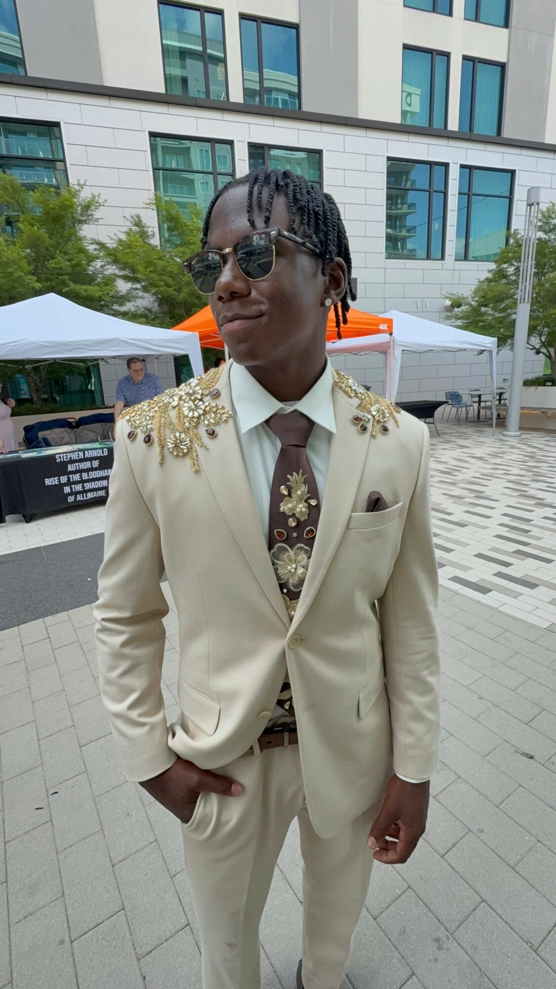 Person in a cream suit with gold embellishments on shoulders, wearing sunglasses, standing outdoors with tents and modern building in background.
