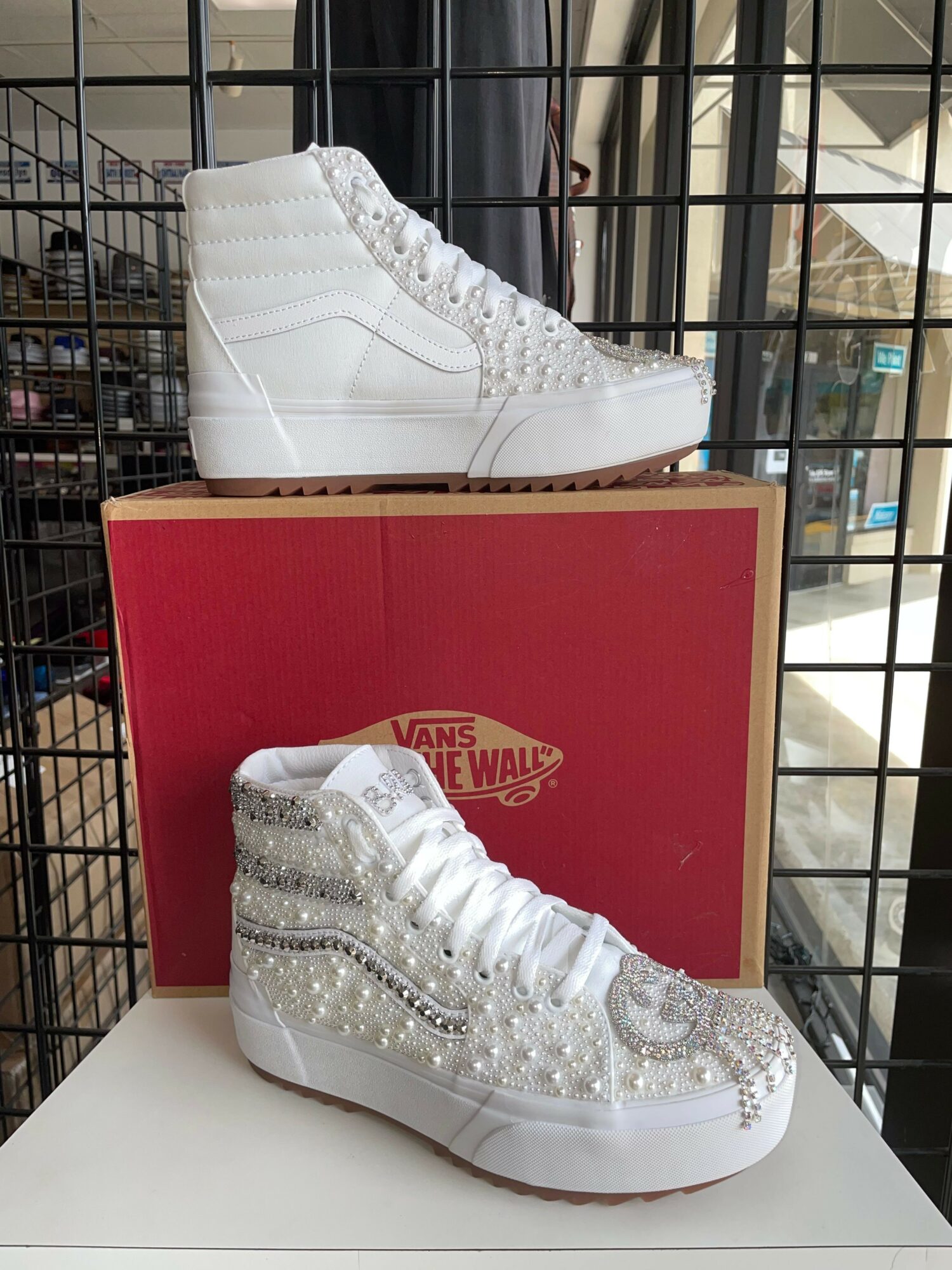 Pair of white high-top sneakers with silver embellishments, placed on a white surface with a red shoe box behind them.