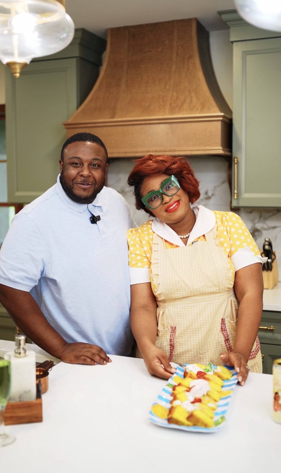 Two people stand in a kitchen with green cabinets and a wooden range hood, smiling at the camera, with a colorful cake on the counter.