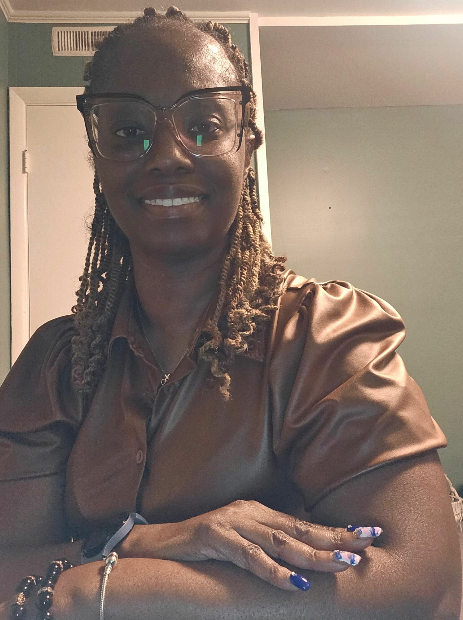 Woman with glasses and long braided hair smiling, wearing a shiny brown top, sitting indoors.
