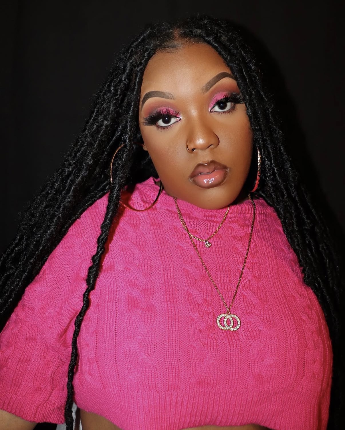 Young woman with long braided hair wearing pink top and gold jewelry, makeup with pink eyeshadow, against dark background.