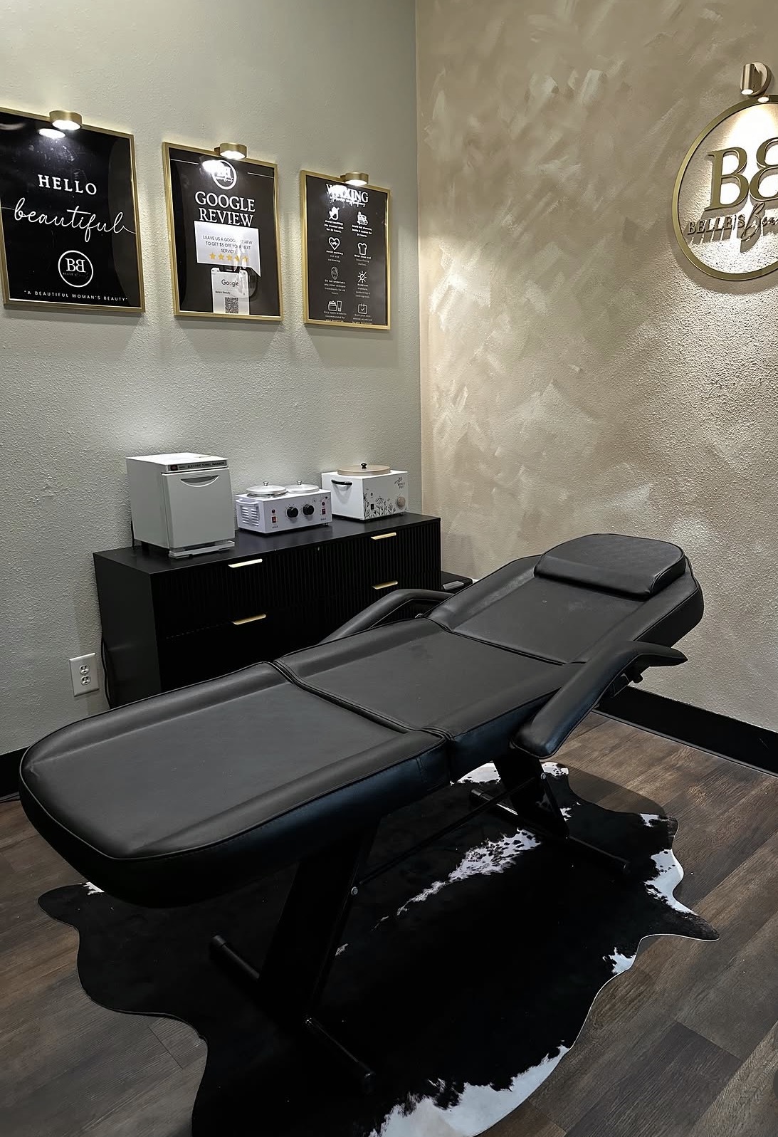 Empty massage or treatment table in a room with framed posters on the wall and a black cabinet, with a black rug underneath.