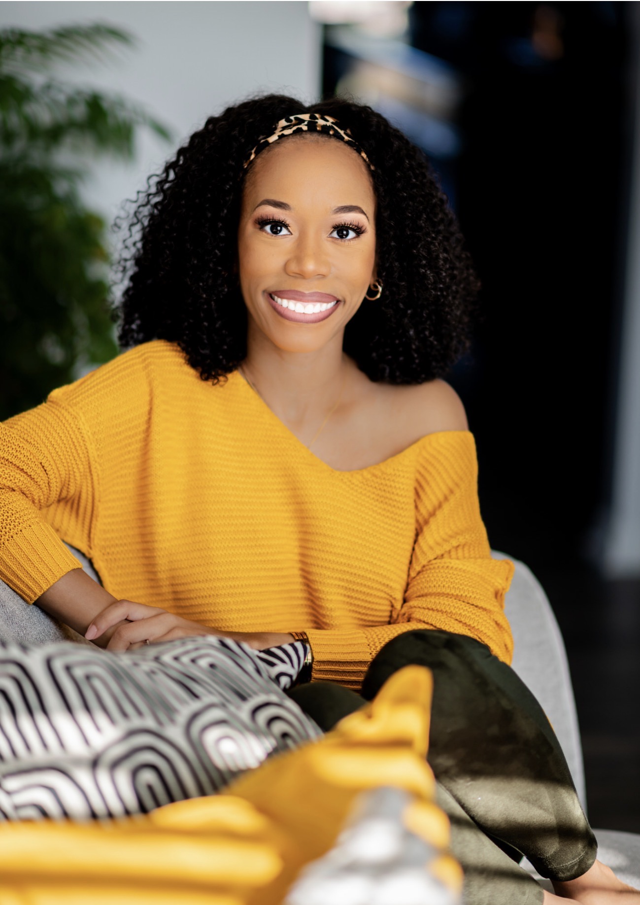 Smiling woman with curly hair wearing a yellow off-shoulder sweater, sitting indoors with a patterned pillow nearby.
