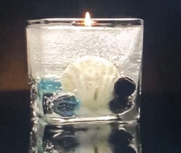 A lit candle in a glass container with seashells and berries inside, reflected on a surface.