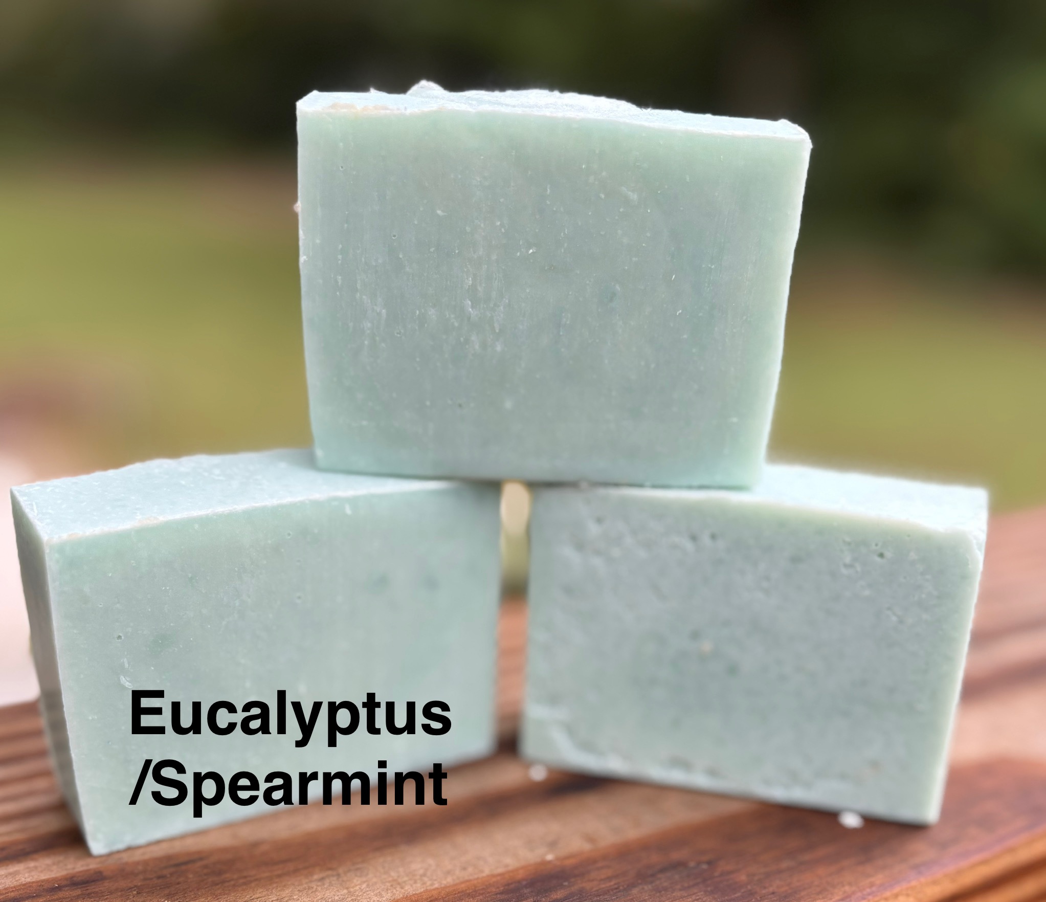 Three light green soap bars stacked outdoors, with one labeled eucalyptus and spearmint, on a wooden surface.