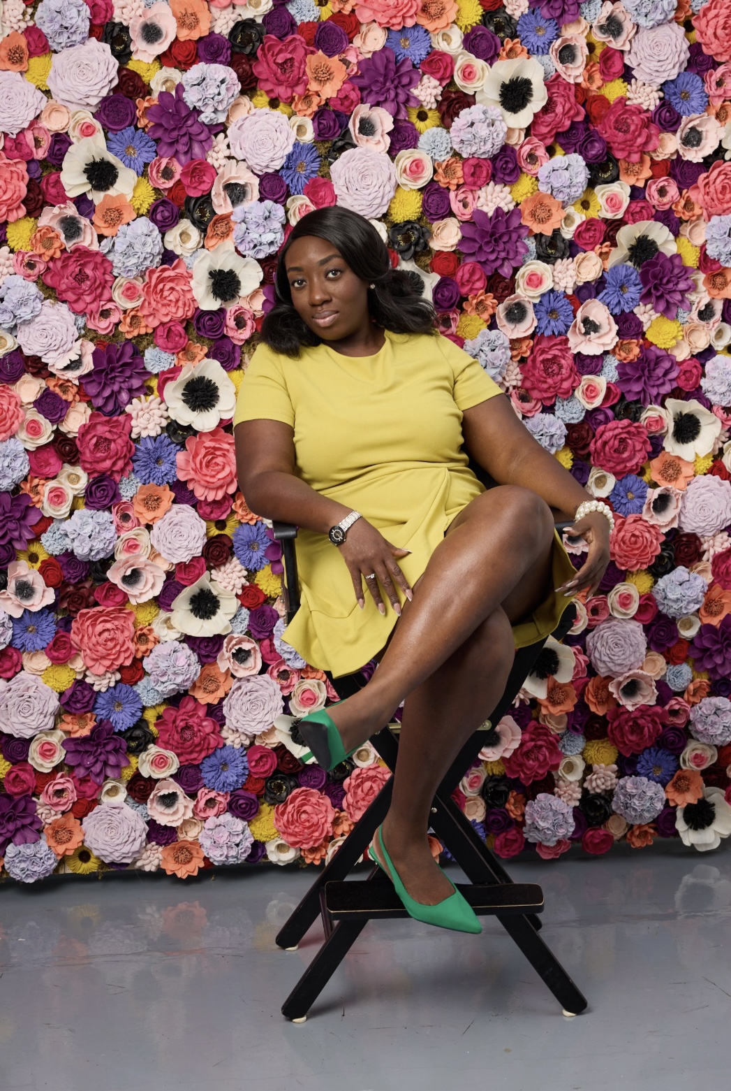 Woman in yellow dress sitting on a stool in front of a colorful flower wall, with crossed legs and green shoes.