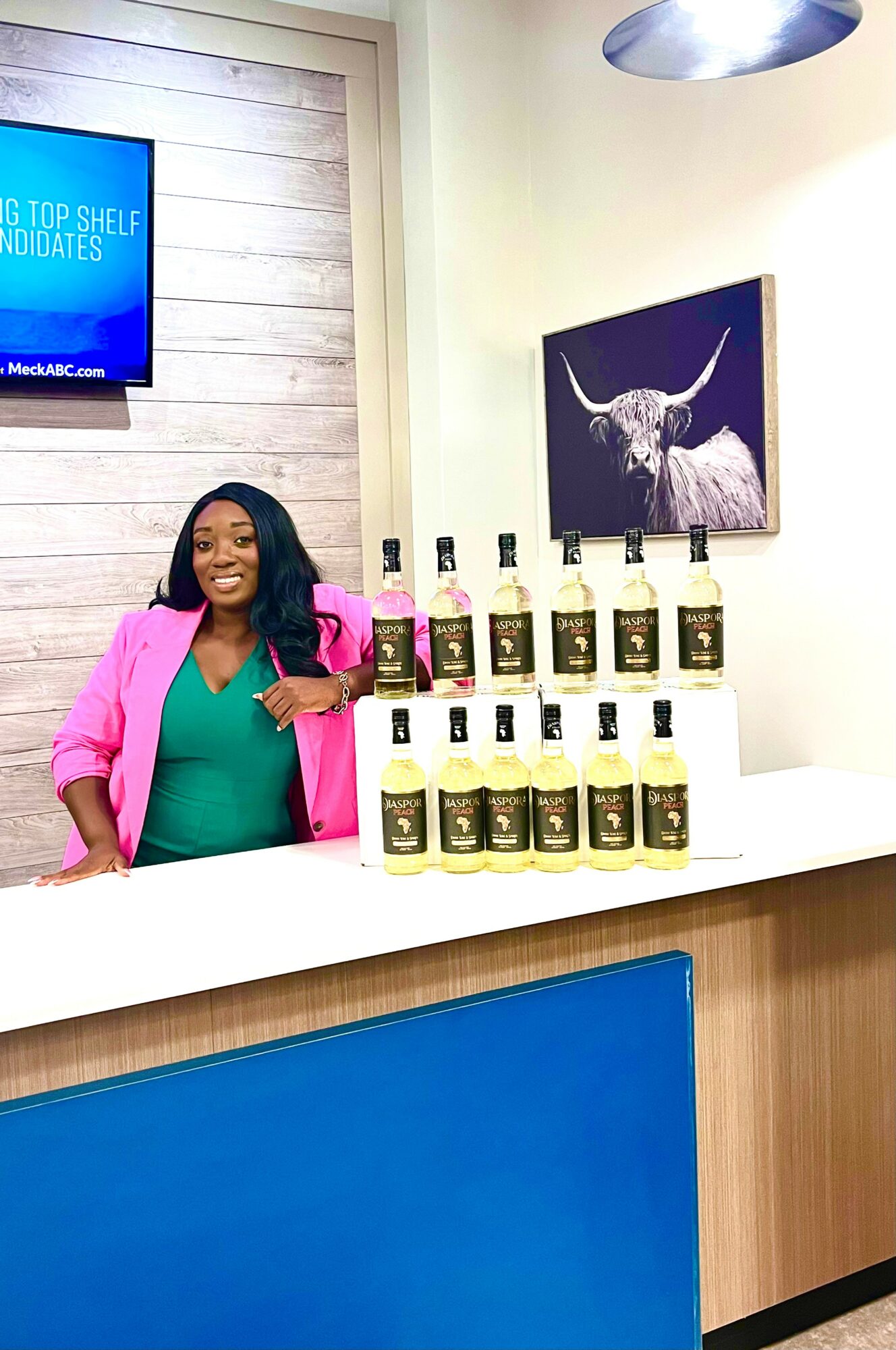 Woman in pink blazer standing behind a counter with bottles, smiling, in a room with a bull painting and a TV screen.
