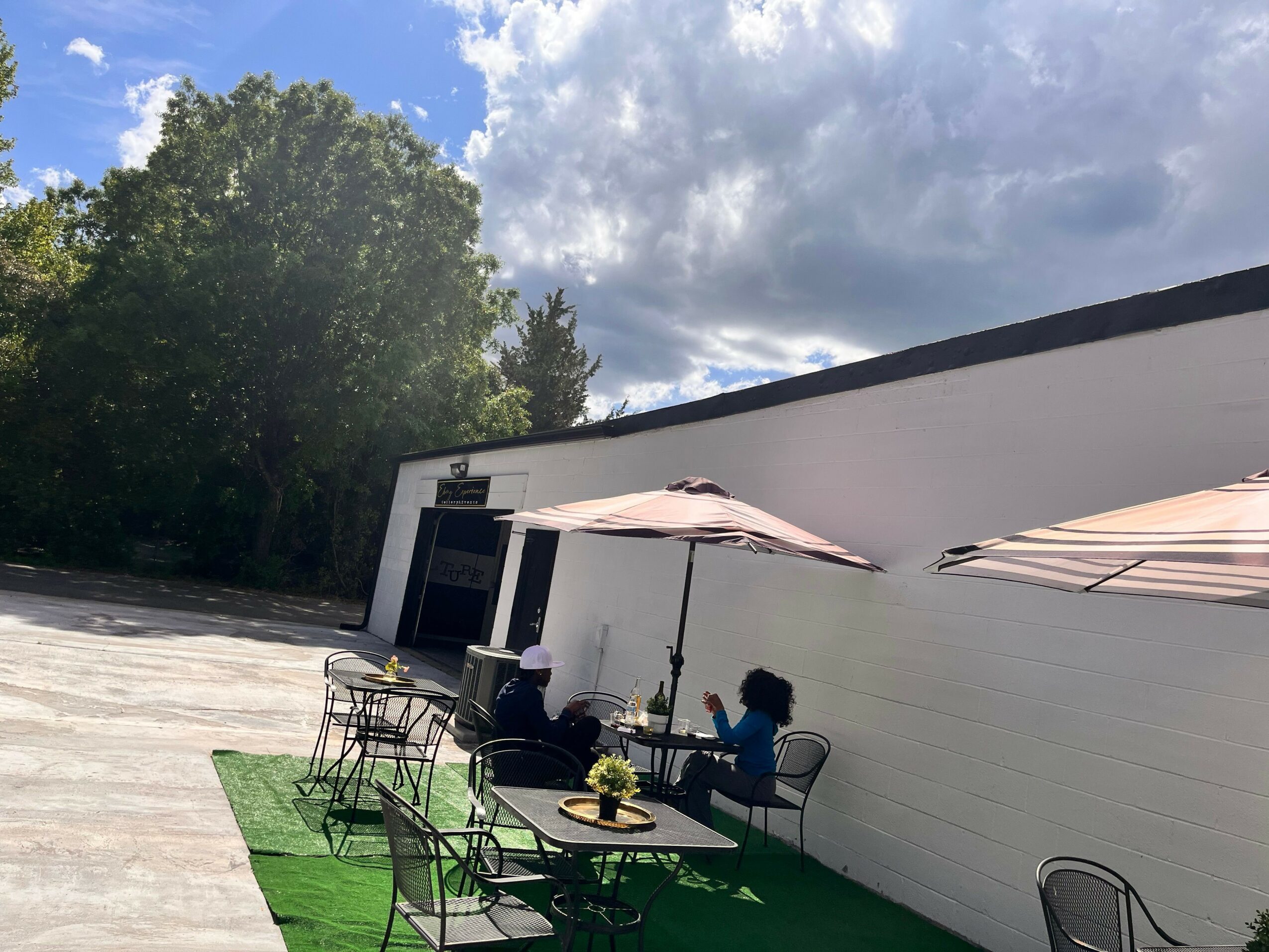 Outdoor patio with green grass, black chairs, and a table, adjacent to a building with a white wall, under a cloudy sky.