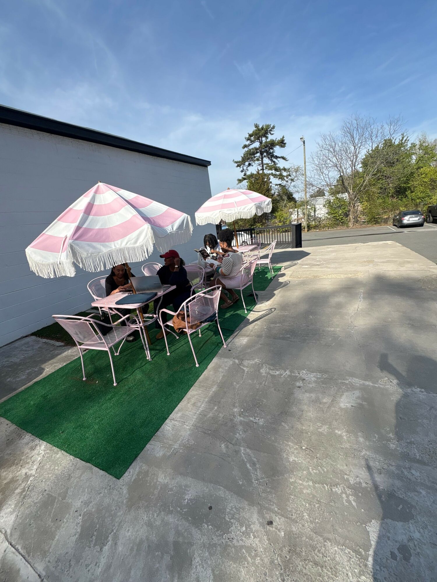 People sitting at tables under umbrellas outside, with a parking lot and trees in the background.