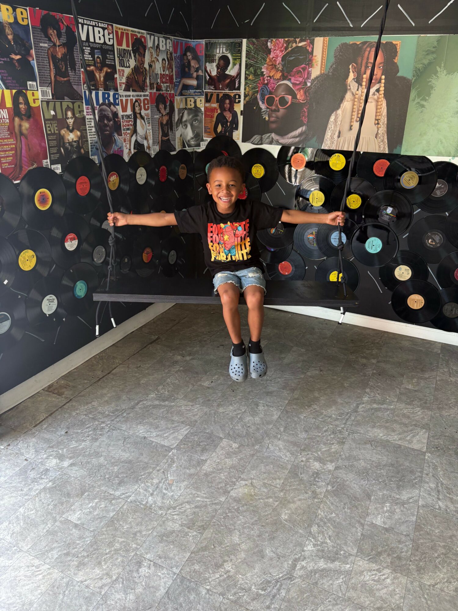 Child standing in front of a wall decorated with colorful circular patterns, holding a drink, with a gray floor.