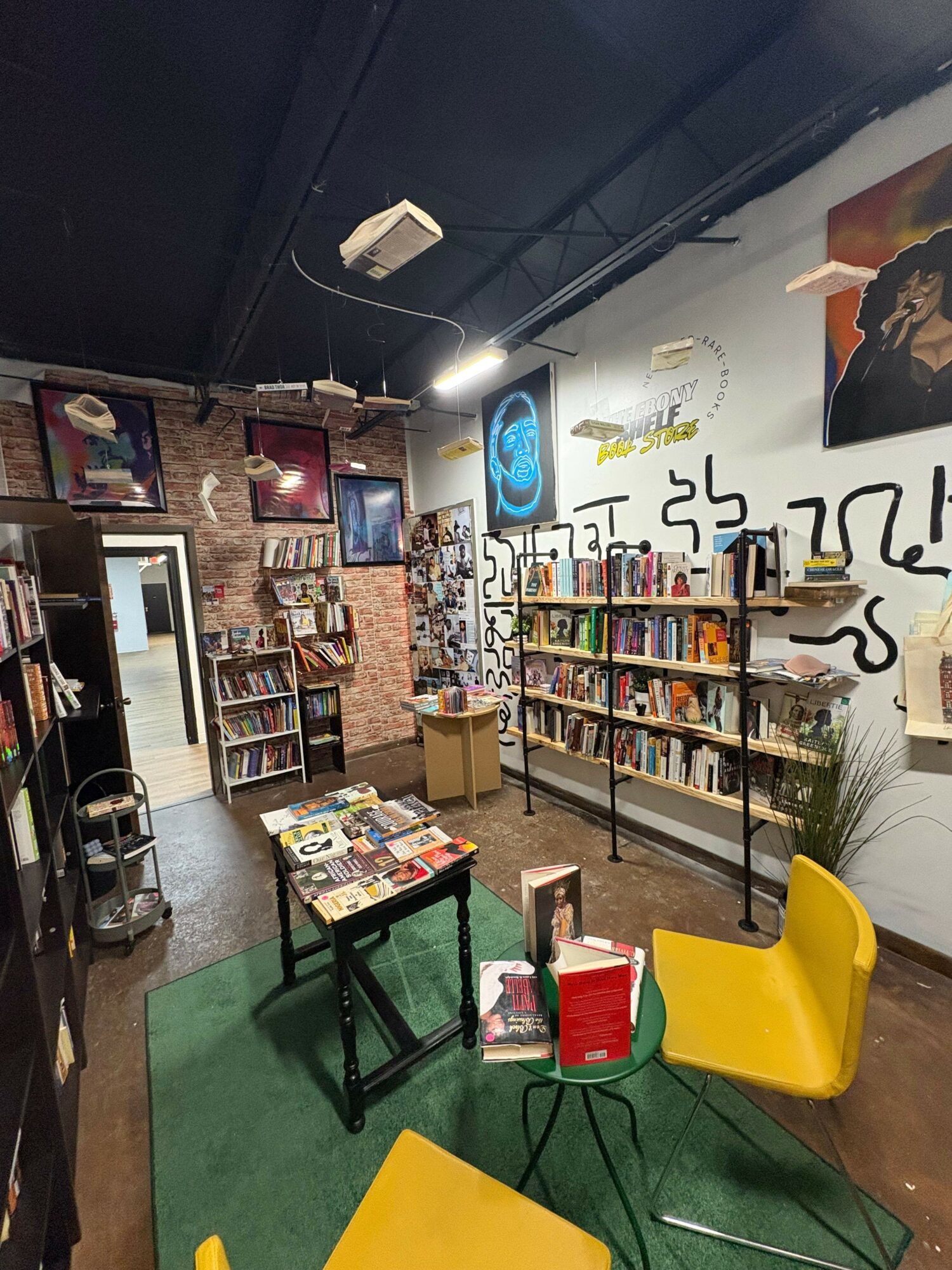 Bookshelves filled with books, a table with magazines, yellow chairs, and a brick wall background in a cozy space.